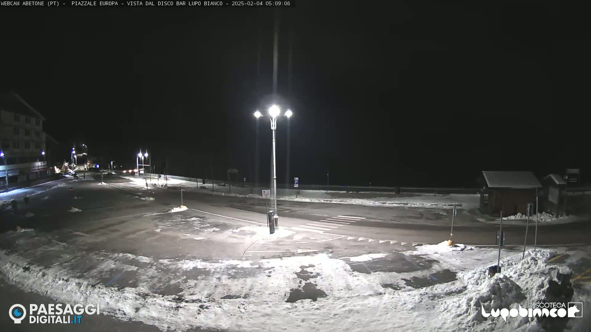 A mostly empty, snow-dusted parking lot is illuminated by streetlights at night.