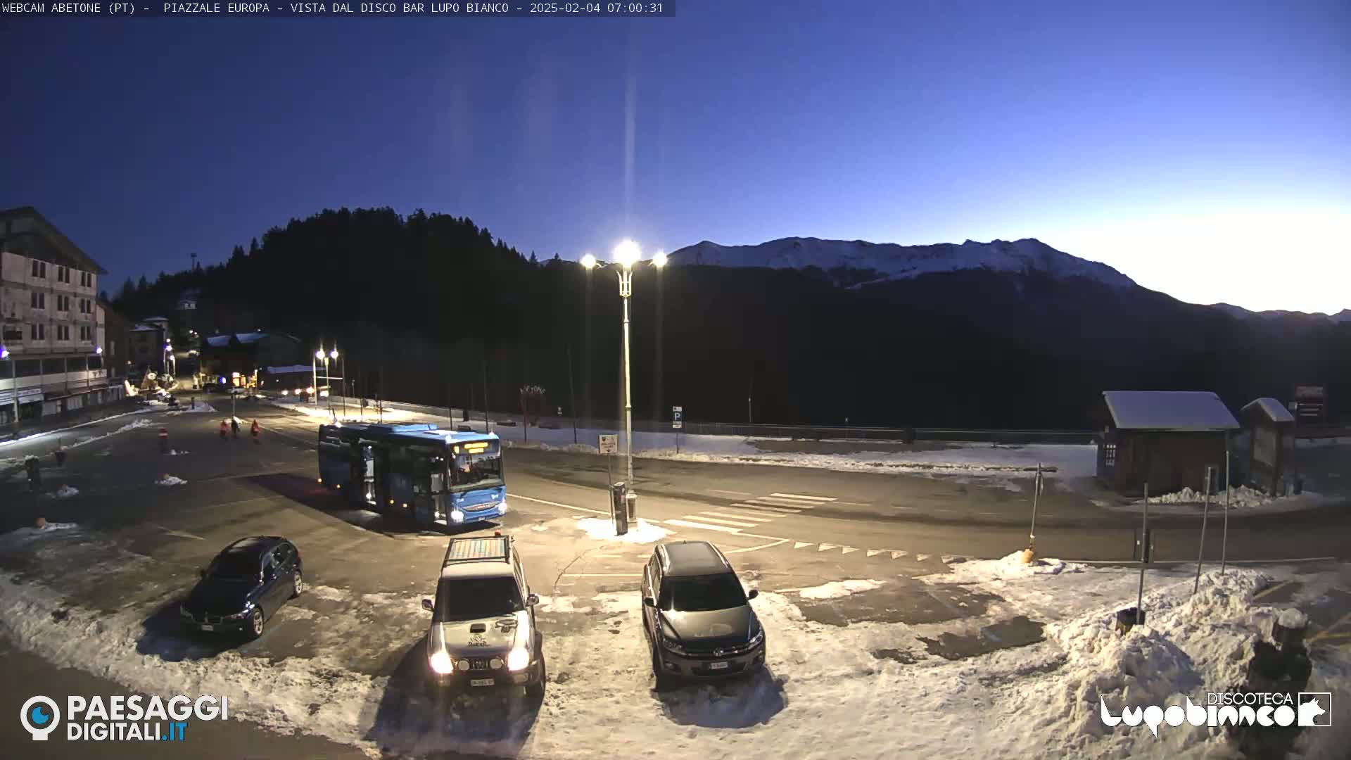 A mostly empty parking lot with a few cars and a bus, situated in front of a building and mountains in the background, under a twilight sky with patches of snow on the ground.