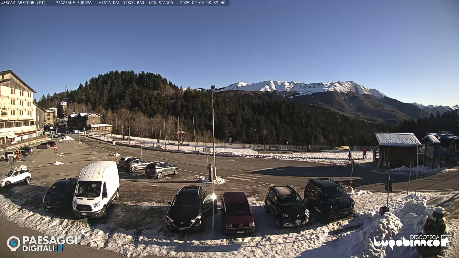 A parking lot with several cars and a van is situated in front of buildings and a snow-capped mountain range under a clear, sunny sky.