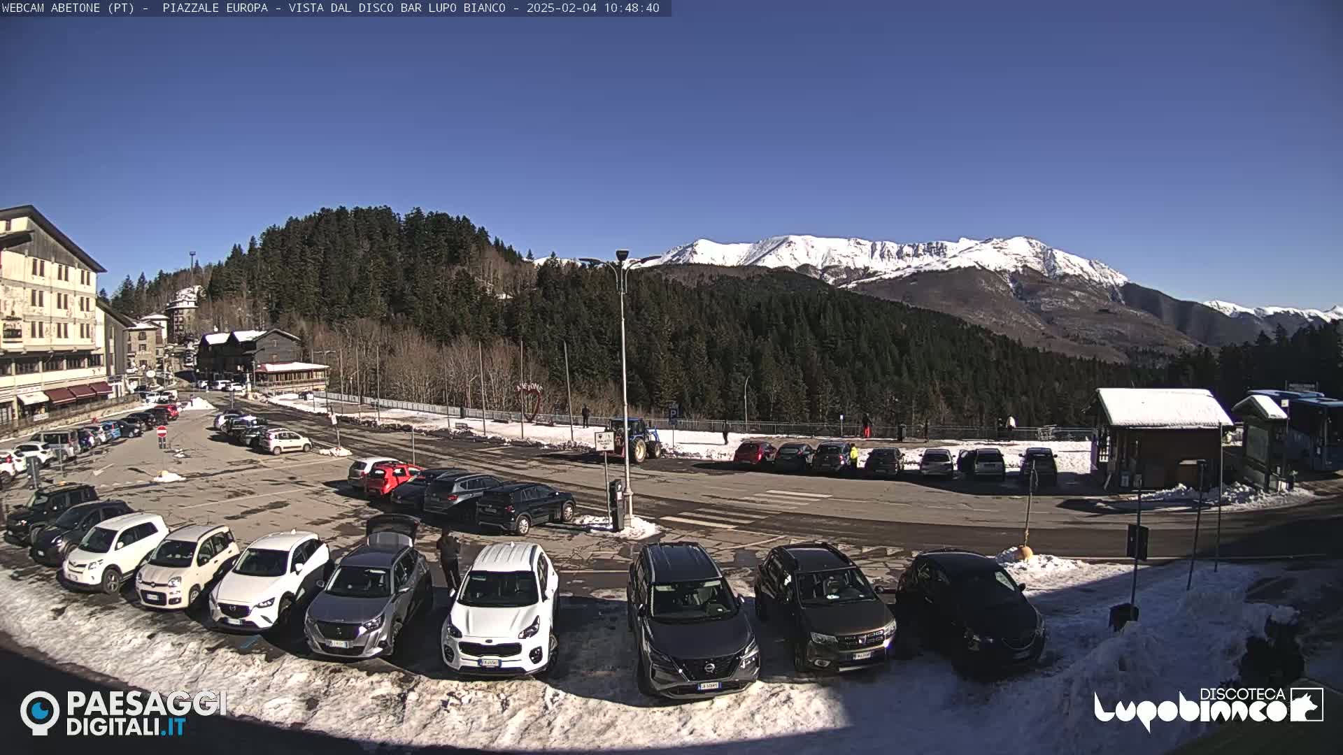 A sunny day in a mountain town parking lot with snow on the ground and snow-capped mountains in the background.