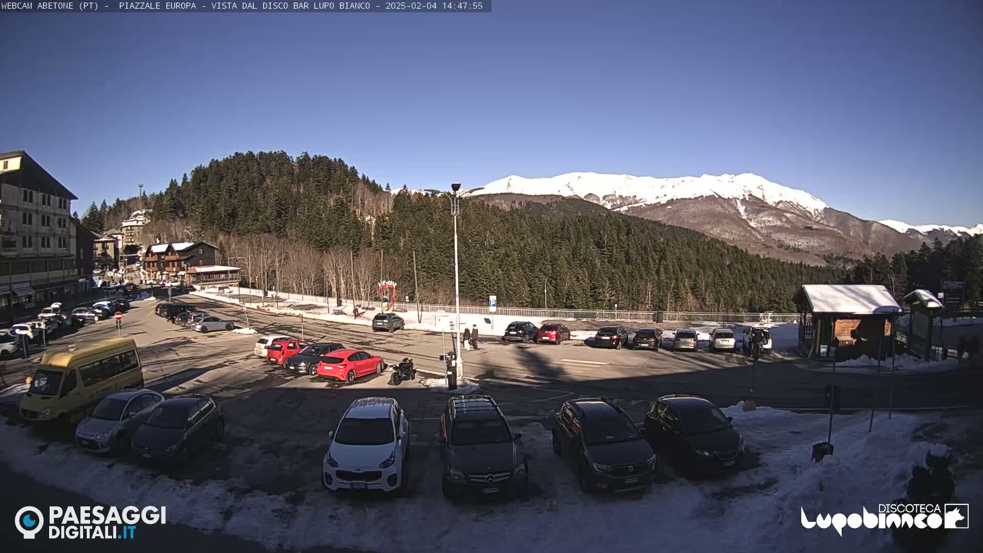A sunny day in a mountain town parking lot, with snow-capped mountains in the background and several cars parked.