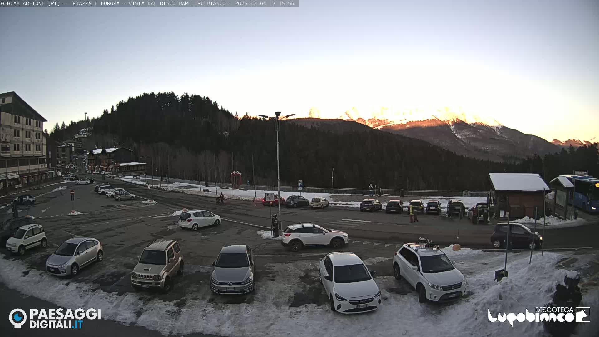 A parking lot with numerous cars is situated in front of buildings, overlooking a snow-dusted landscape with mountains in the background during a clear sunset.