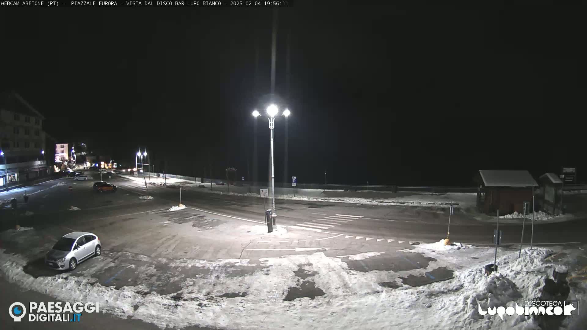 A snow-covered nighttime parking lot with a few cars, streetlights, and buildings in the background.