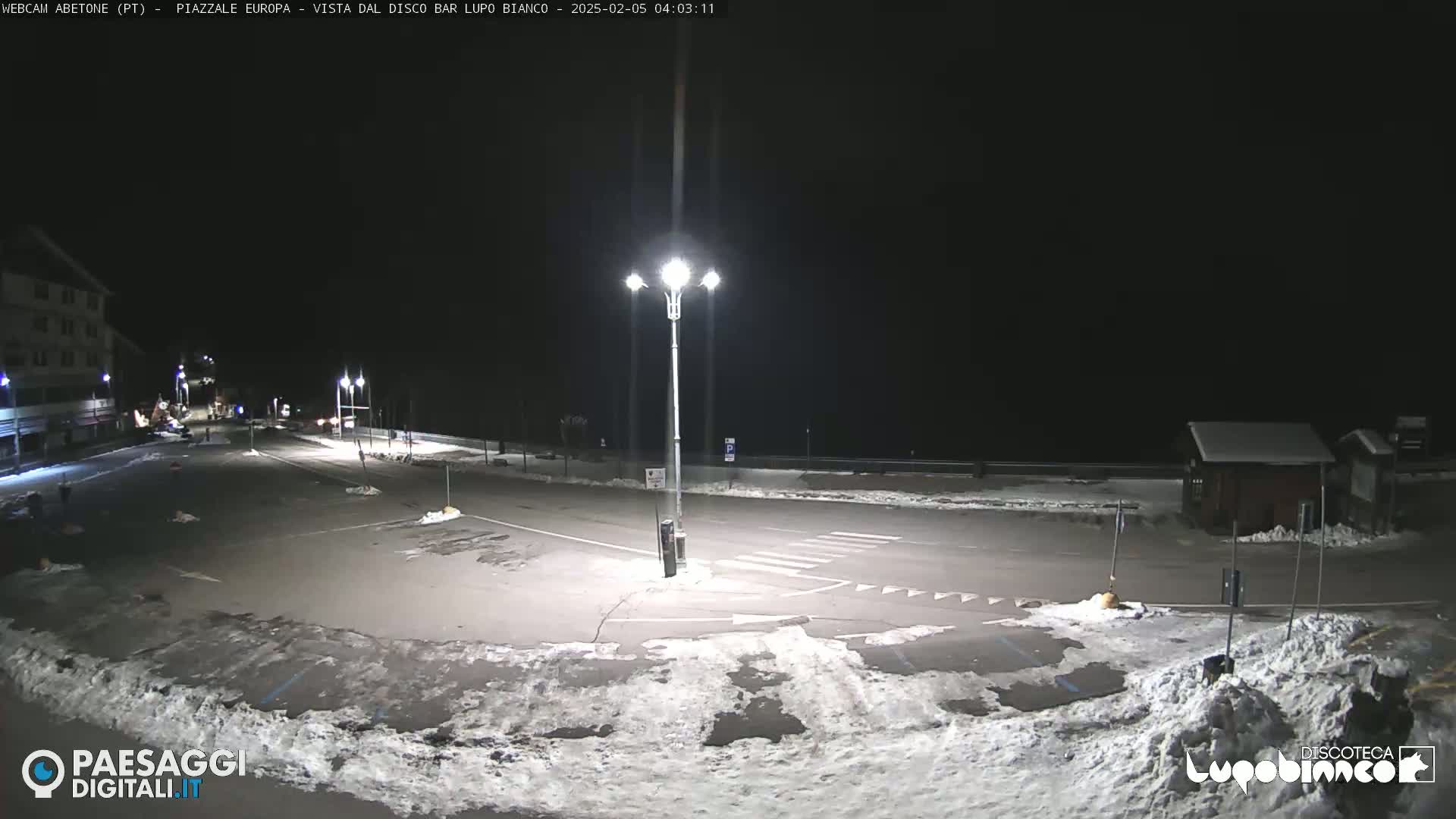 A snow-covered, empty parking lot is illuminated by streetlights at night.
