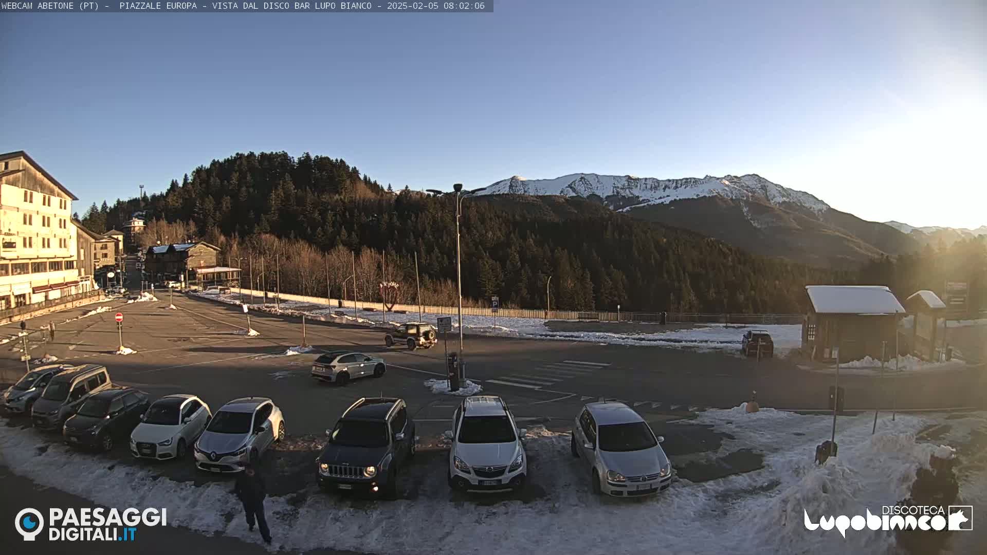 A parking lot with several cars is situated in front of a building and a snow-covered mountain range under a clear, bright sky.