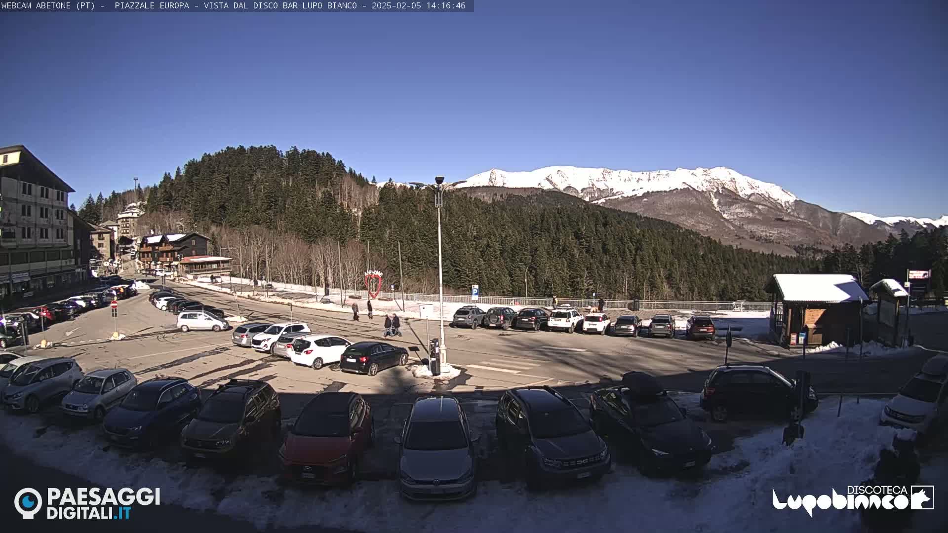 A sunny day in a parking lot, surrounded by snow-capped mountains and evergreen trees.
