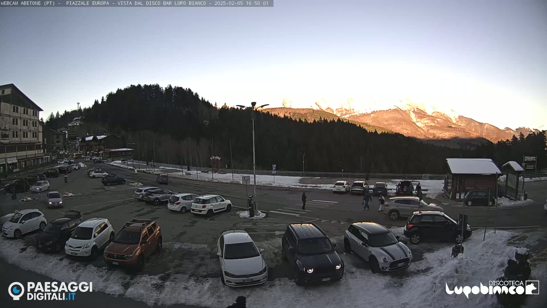 A parking lot filled with cars is situated in front of a mountain range at sunset, with some snow visible on the ground.