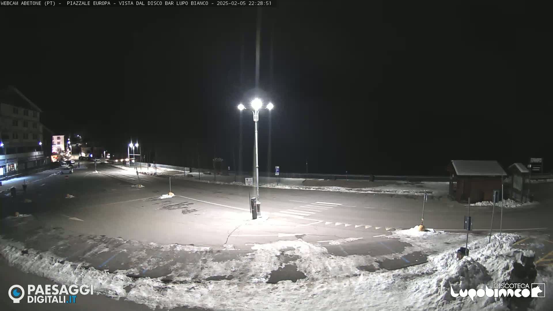 A mostly empty nighttime parking lot with snow on the ground and a few buildings visible in the distance under a clear, dark sky.