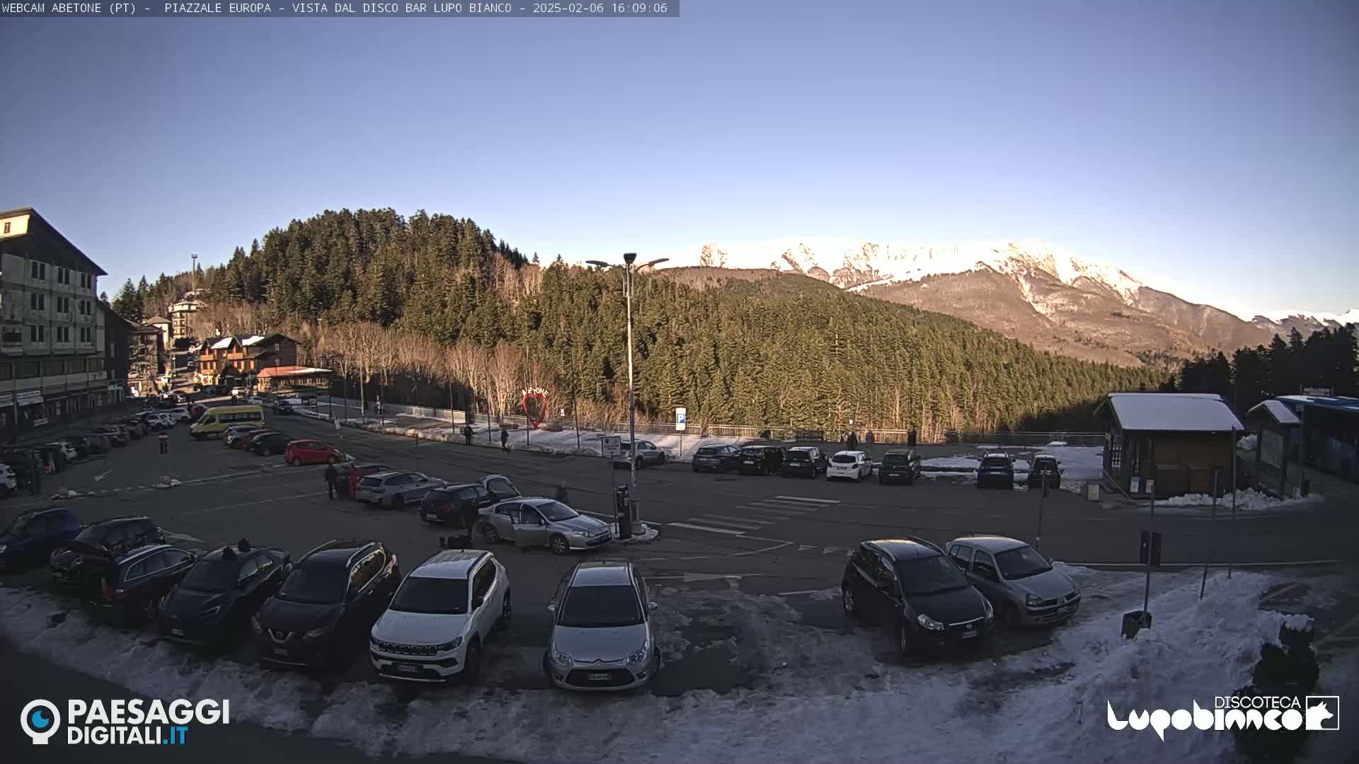 A parking lot with several cars is situated in front of a mountain range covered with evergreen trees, under a clear, sunny sky.