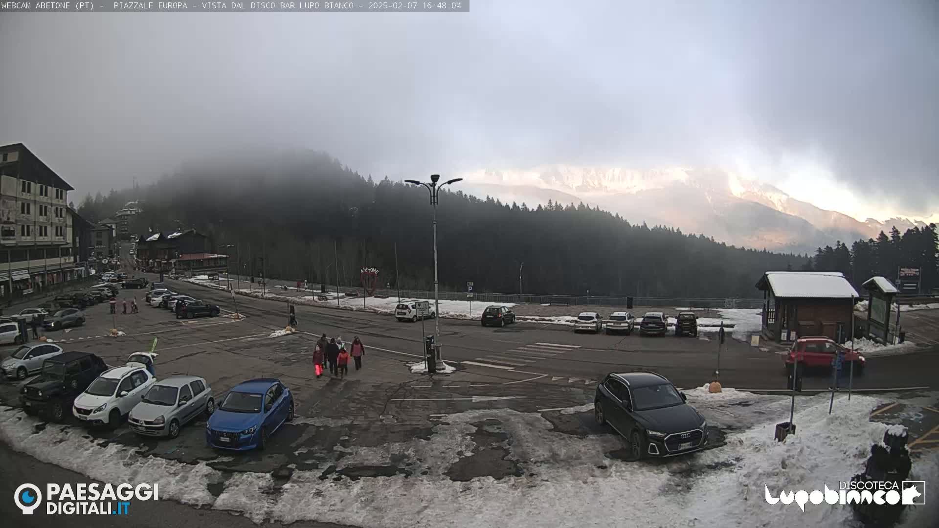 A mostly cloudy day reveals a mountain range in the distance, partially obscured by fog, overlooking a parking lot with several cars and a few pedestrians.