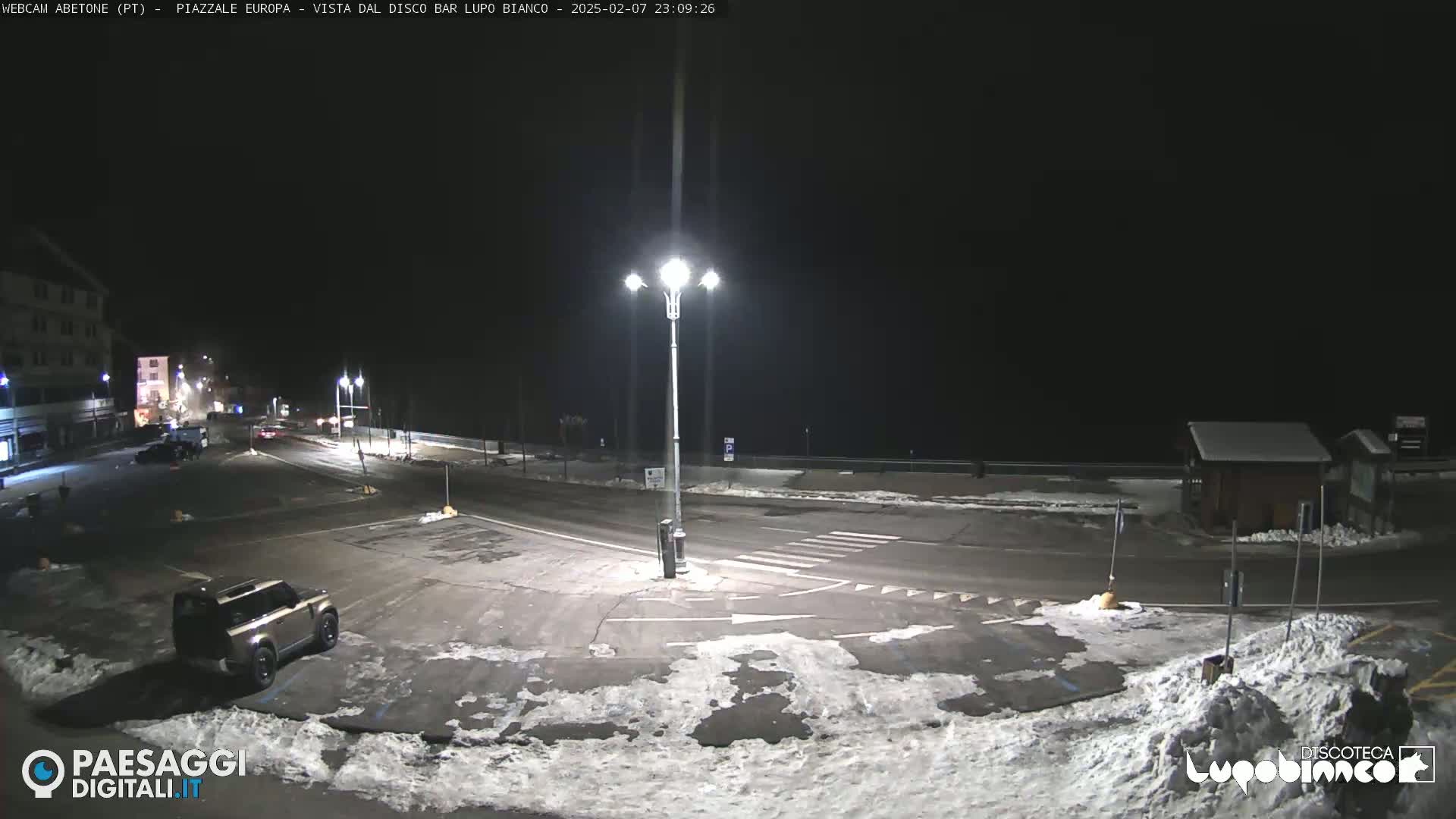 A nighttime view of a snow-covered parking lot and roadway, illuminated by streetlights, with a few vehicles visible.