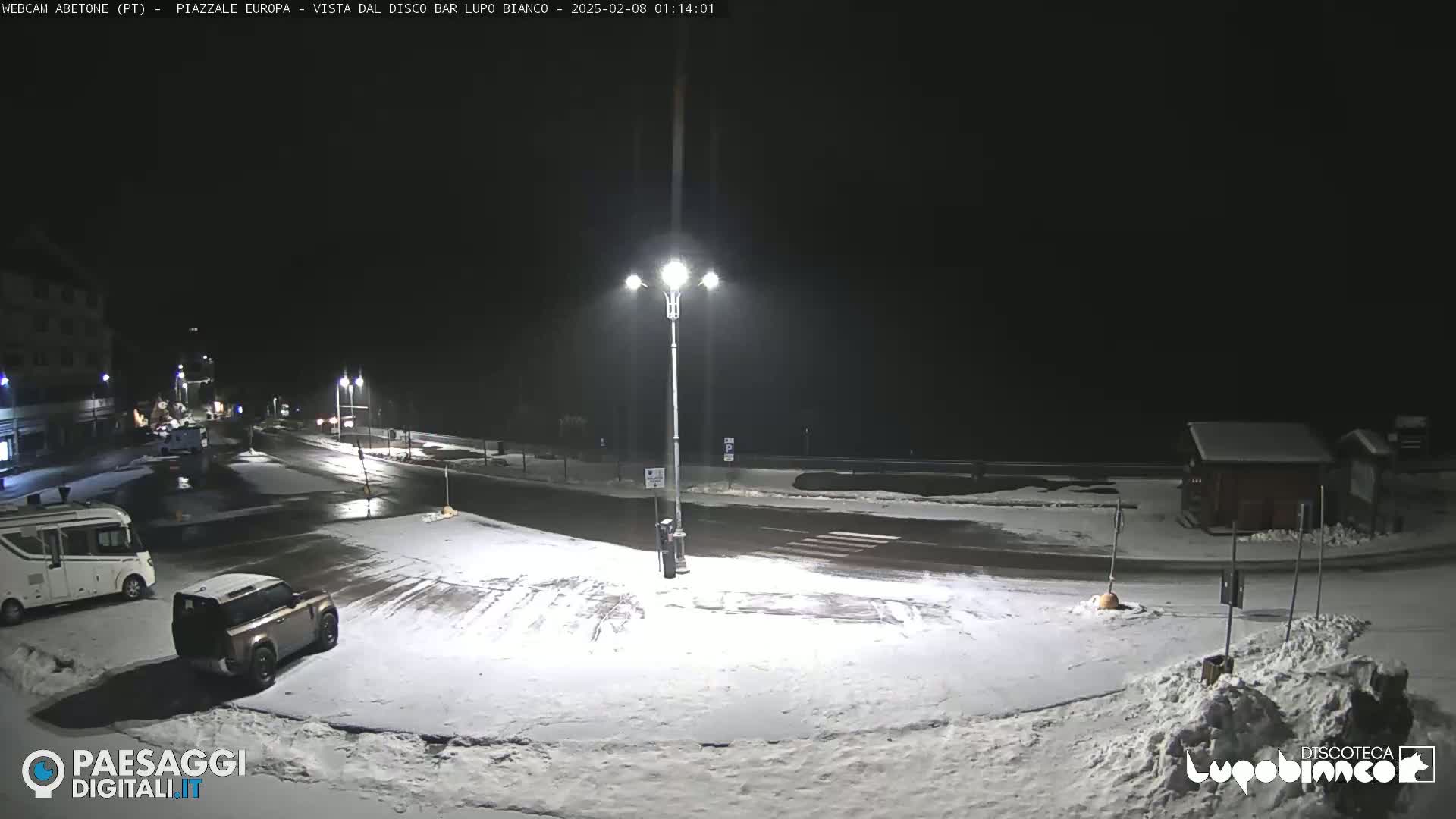 A snow-covered parking lot and road at night, illuminated by streetlights, with a few vehicles visible.