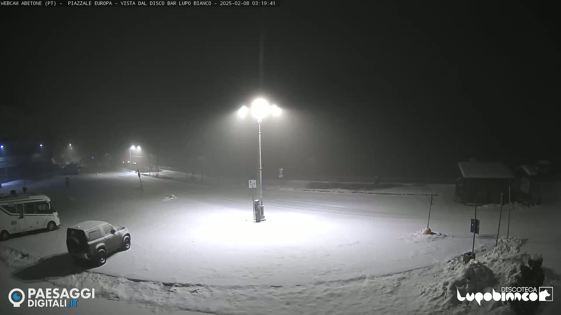 A snow-covered parking lot at night, illuminated by several bright streetlights in foggy conditions, with a few vehicles parked.