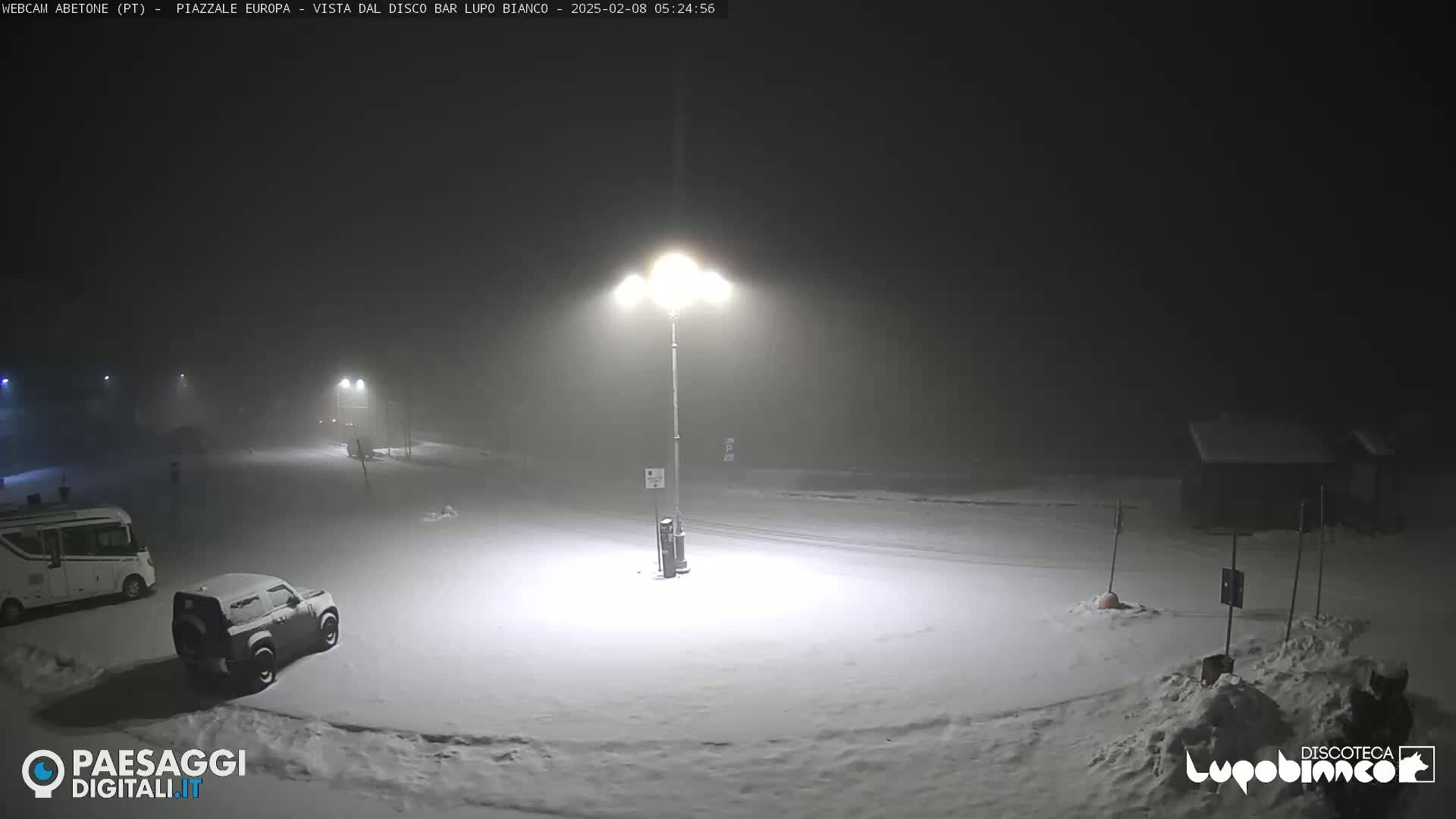 A snow-covered parking lot is dimly lit at night under foggy conditions, with a few vehicles visible.