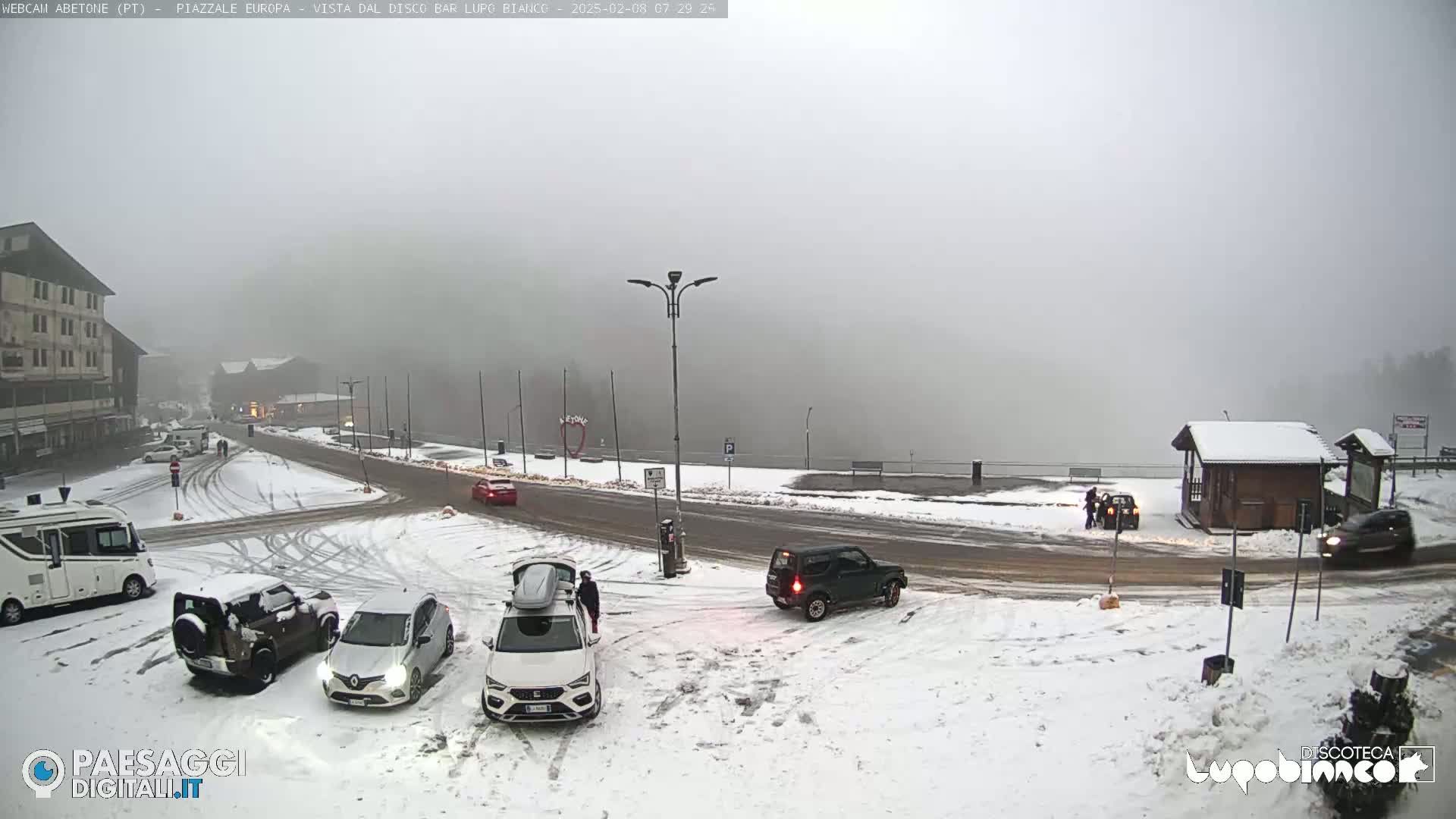 A snow-covered parking lot and road are visible in foggy conditions, with several cars and a camper van parked amidst the snow.