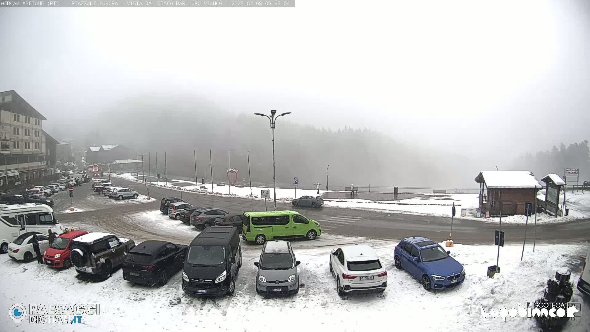 A snow-covered parking lot with cars and a building in the background, all under a foggy sky.