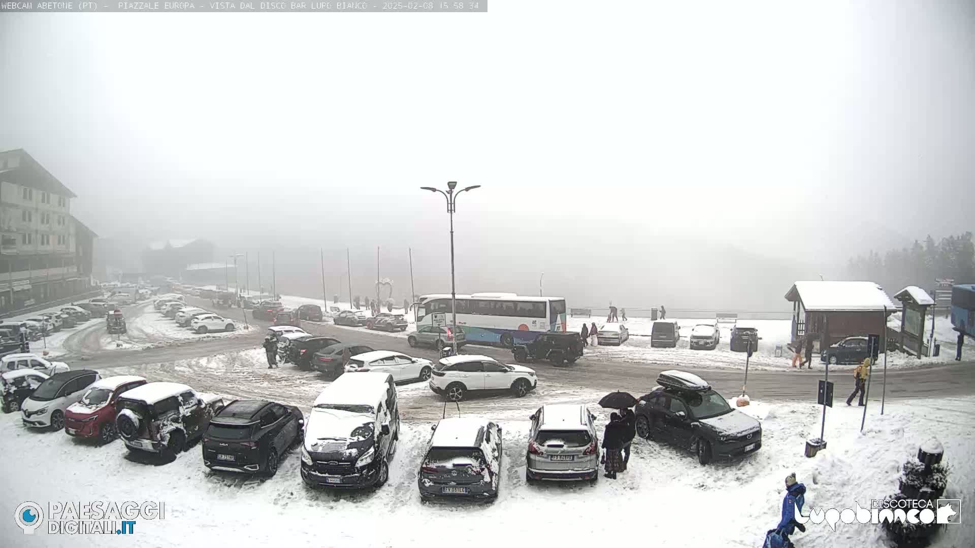 A snow-covered parking lot with several cars and a bus, in foggy weather conditions.