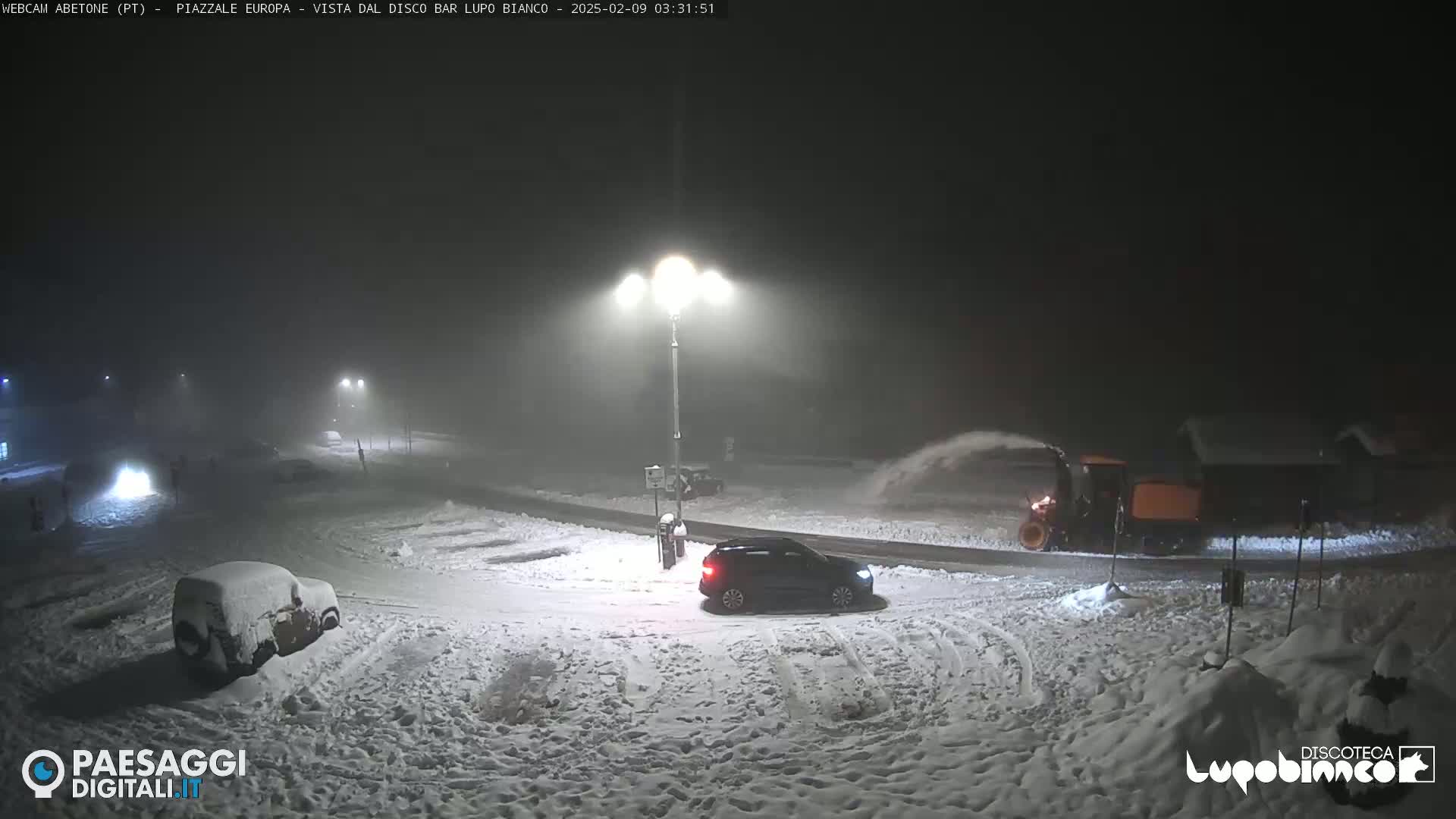 A snowplow clears snow from a snow-covered parking lot at night under dim streetlights.