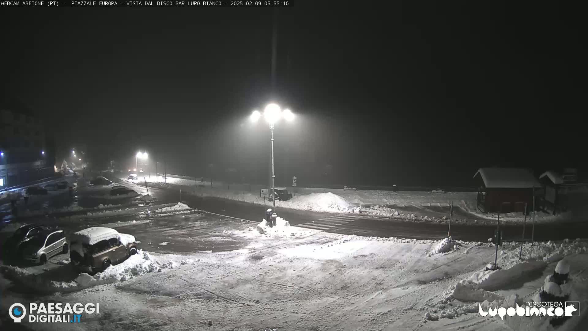 A snow-covered parking lot and road are dimly lit at night under foggy conditions.