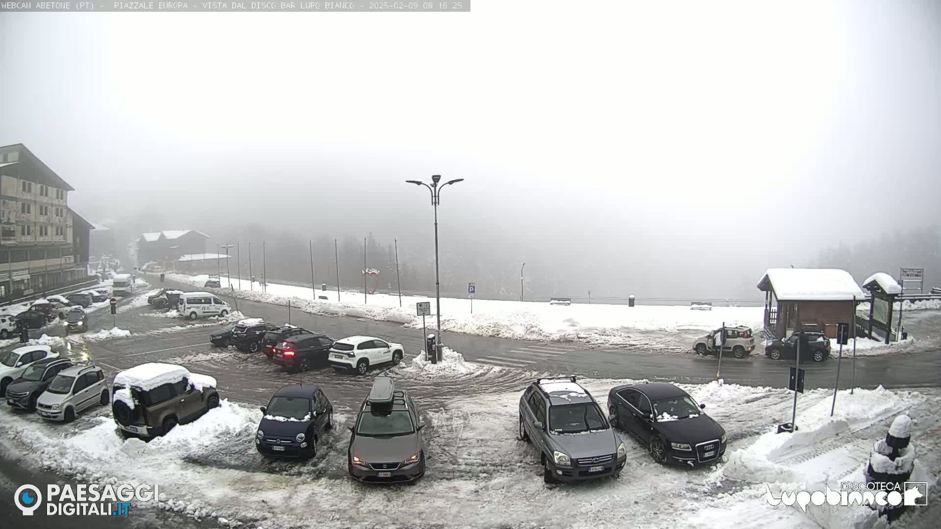 A snow-covered parking lot with several cars, surrounded by snow-covered buildings and roads in foggy conditions.