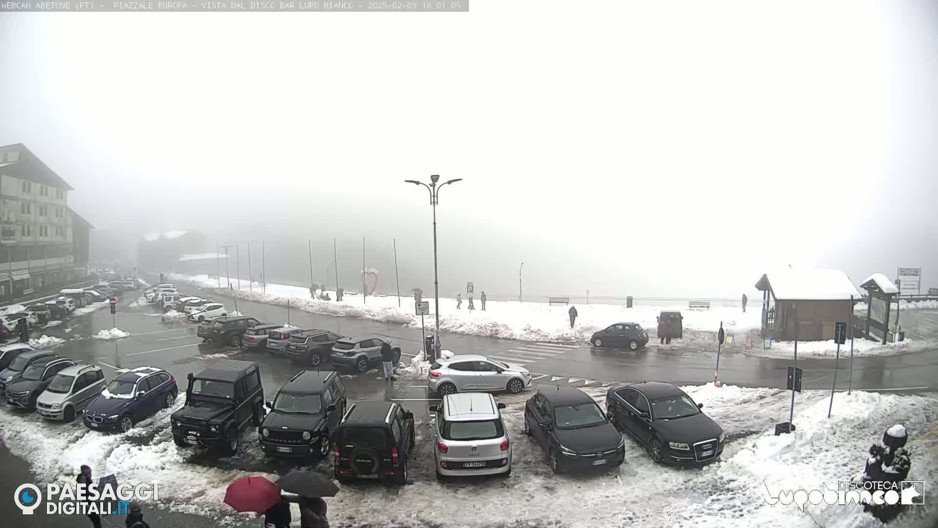 A foggy, snowy parking lot with numerous cars and a few pedestrians, situated near a building.