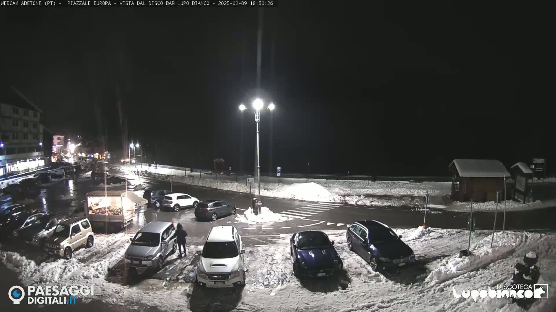 A nighttime parking lot with several cars parked in the snow, illuminated by streetlights.