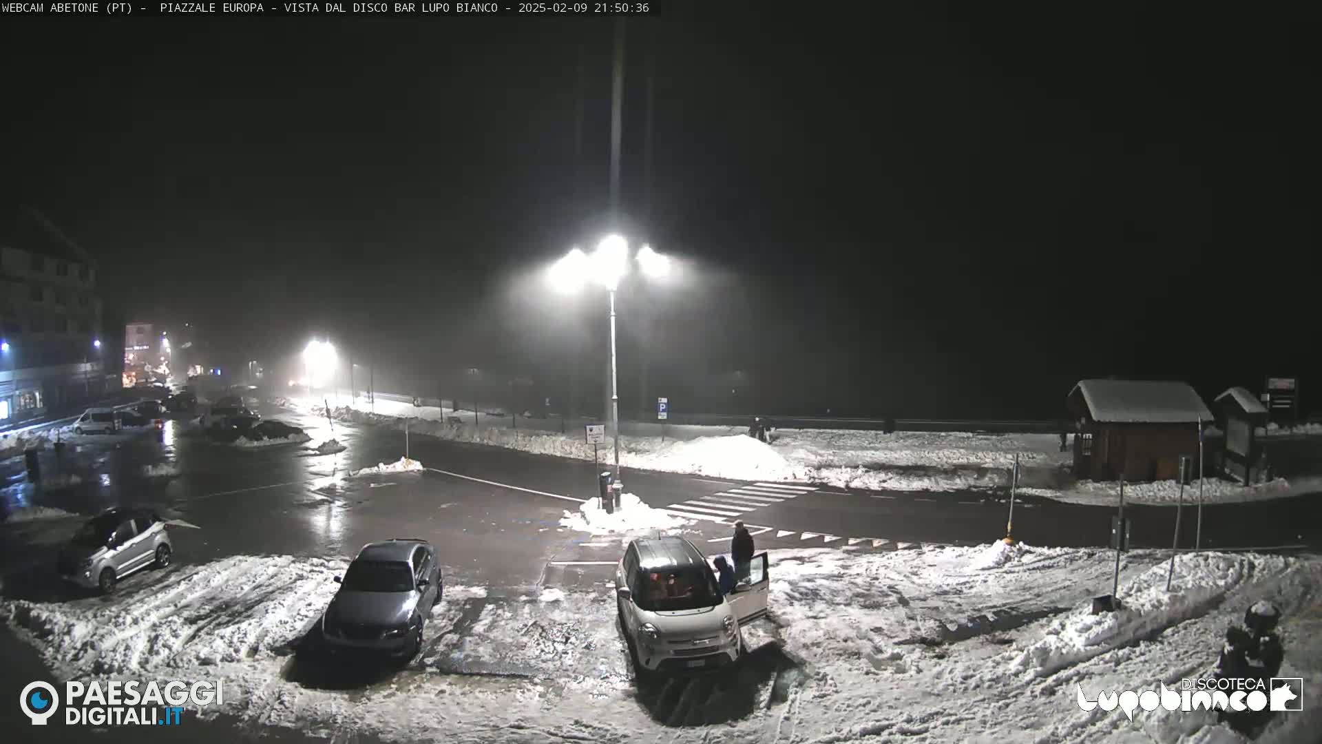 A nighttime view of a snow-covered parking lot with several cars and a few people, under hazy, dimly lit conditions.