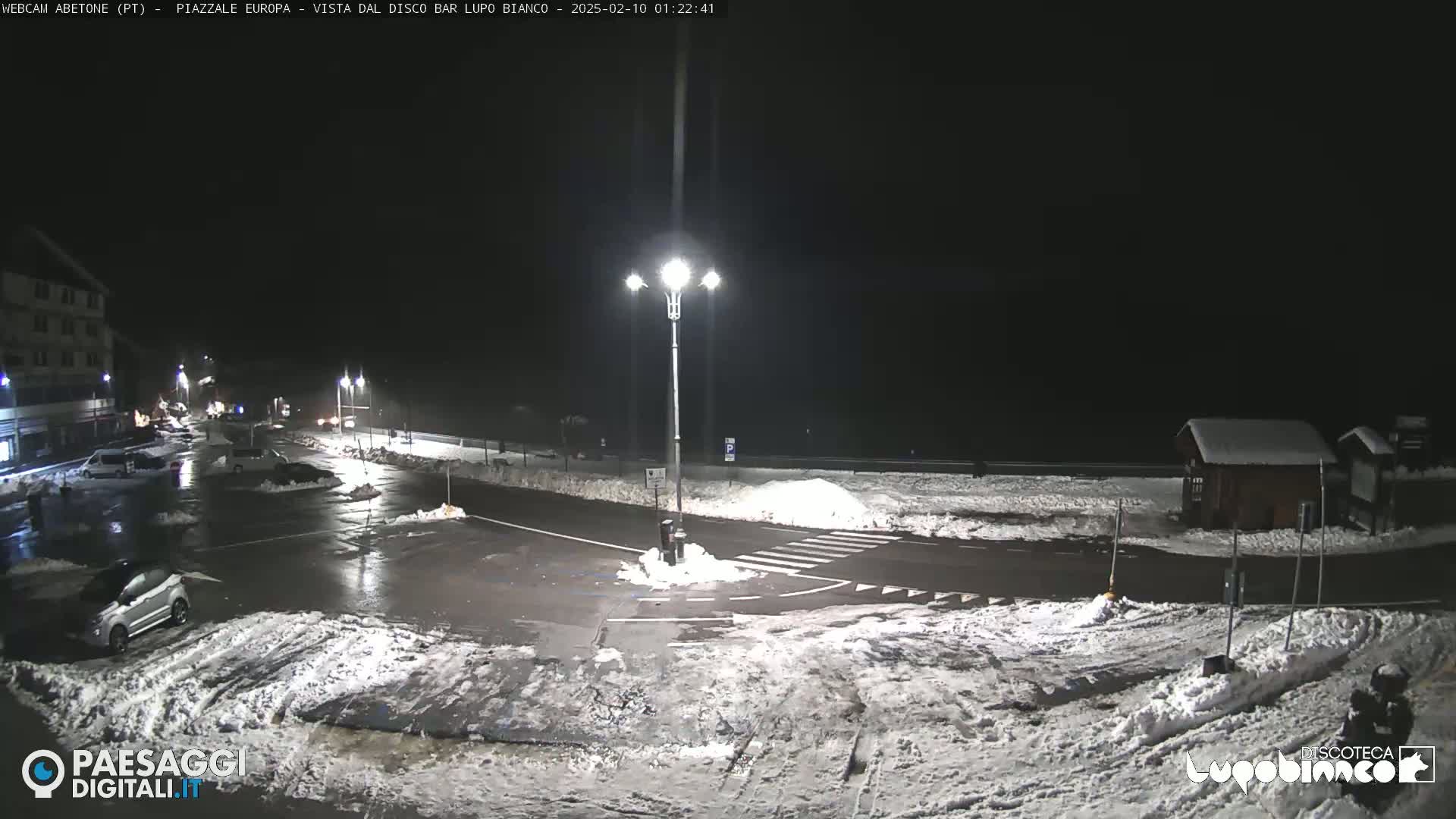 A nighttime view of a snow-covered parking lot and roadway intersection, illuminated by streetlights.