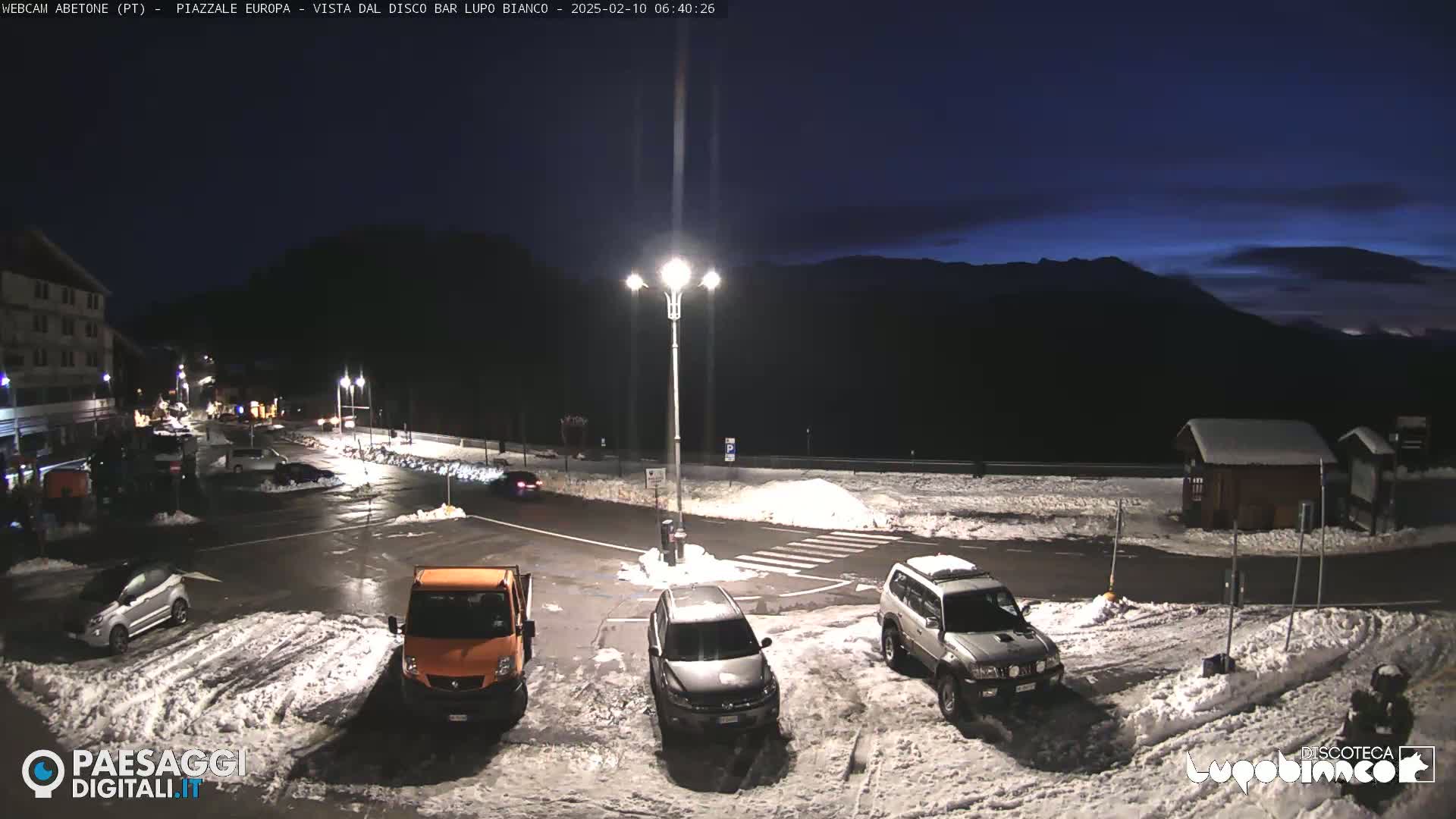 A snow-covered parking lot with several vehicles is illuminated at night under a dark blue twilight sky, mountains visible in the background.