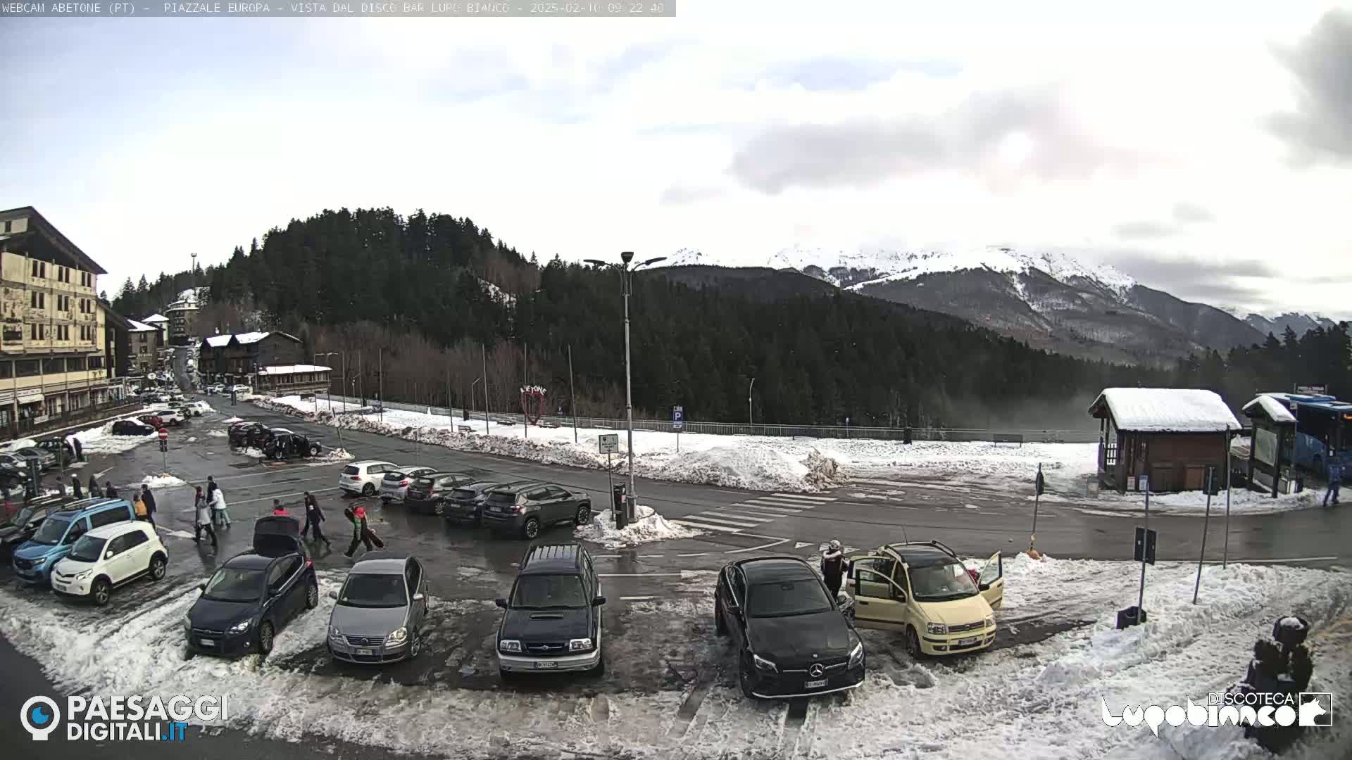 A parking lot with cars and people, partially covered in snow, is situated in front of a building and forested mountains under an overcast sky.