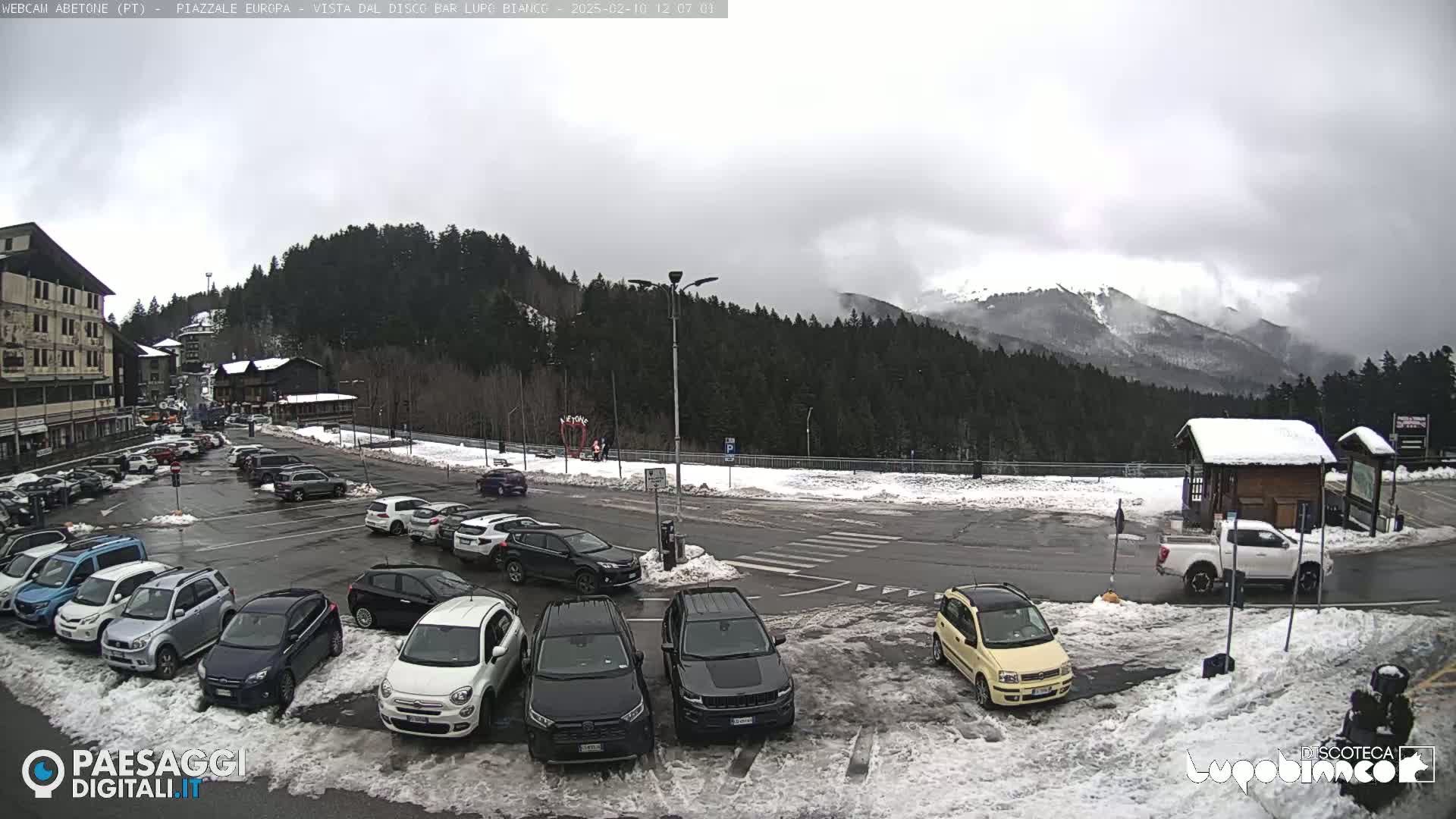 A parking lot with several cars is situated in front of a building, next to a snow-covered landscape with evergreen trees and mountains in the background under an overcast sky.