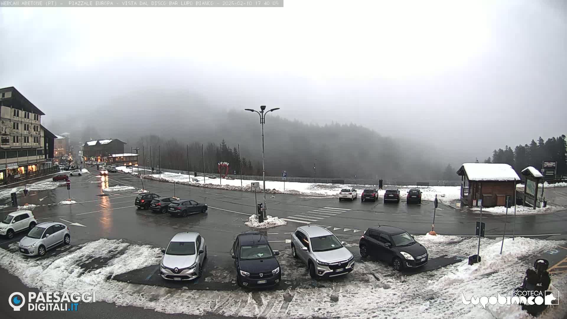 A foggy, snowy parking lot with several cars and a building in the background.