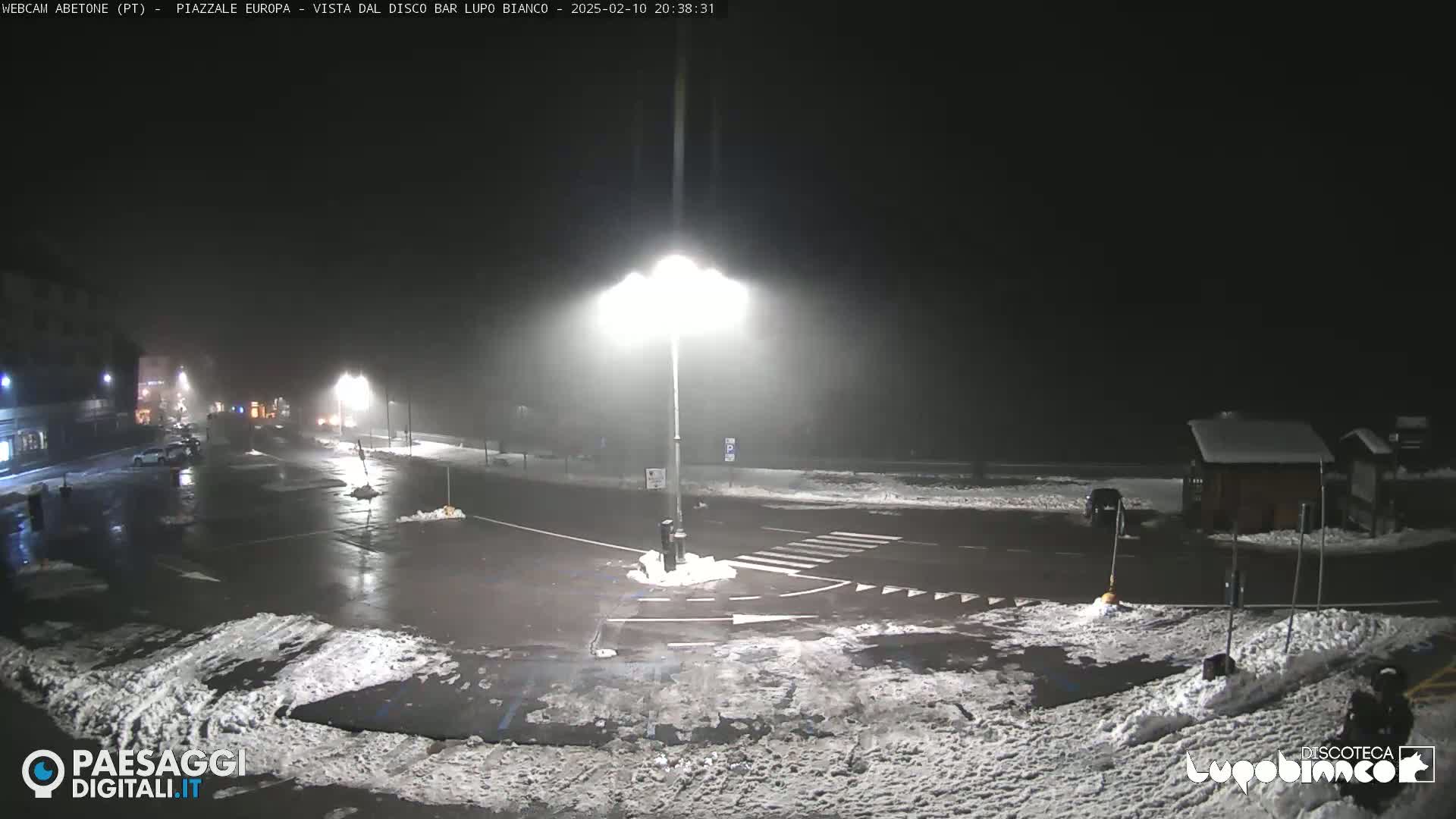 A nighttime, snow-dusted parking lot and roadway intersection are illuminated by bright streetlights under foggy conditions.