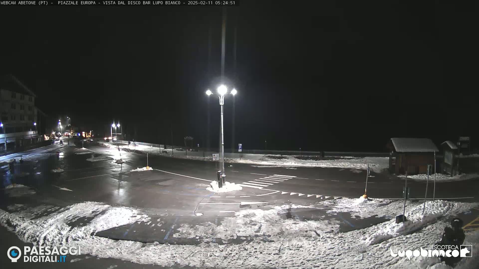 A snow-dusted nighttime parking lot with a street and building in the background under bright artificial lights.