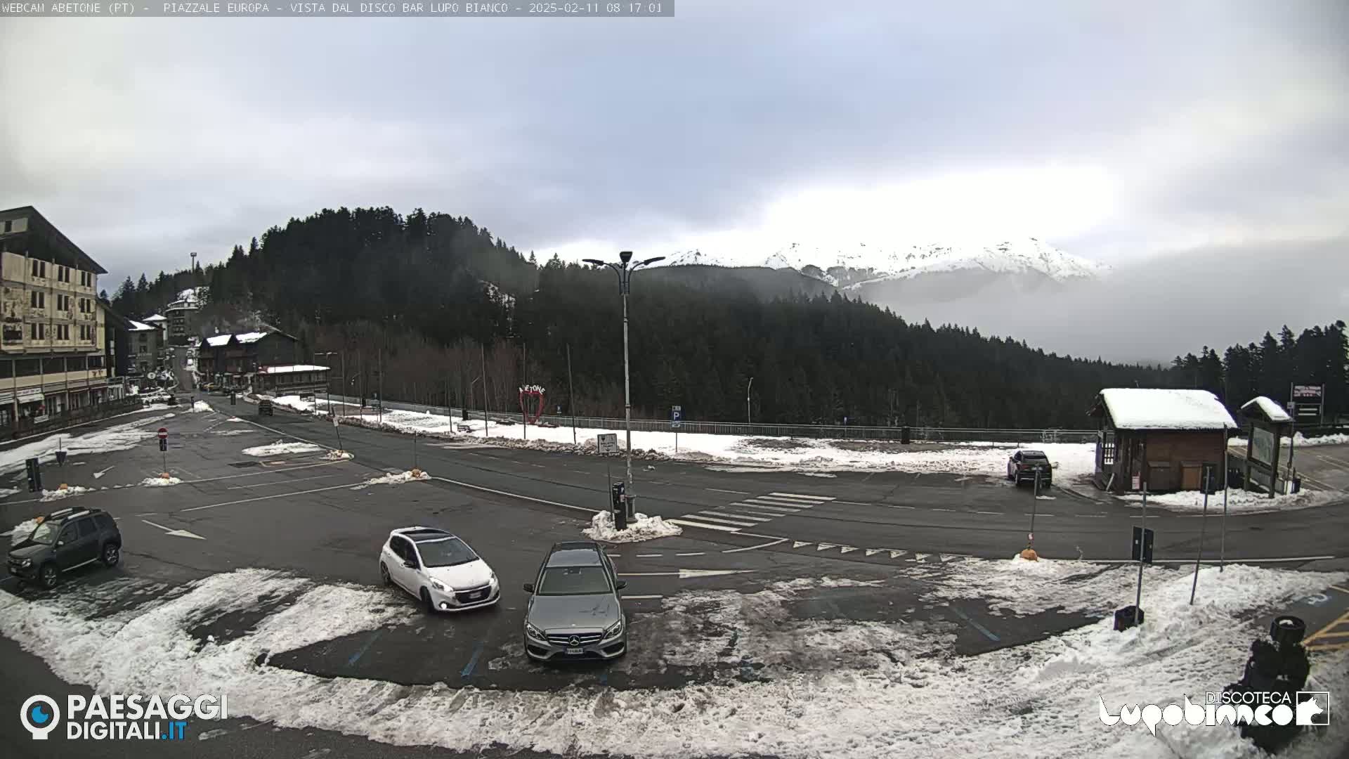 A snow-dusted parking lot with several cars is situated in front of buildings and a snow-covered mountain range partially obscured by fog under an overcast sky.