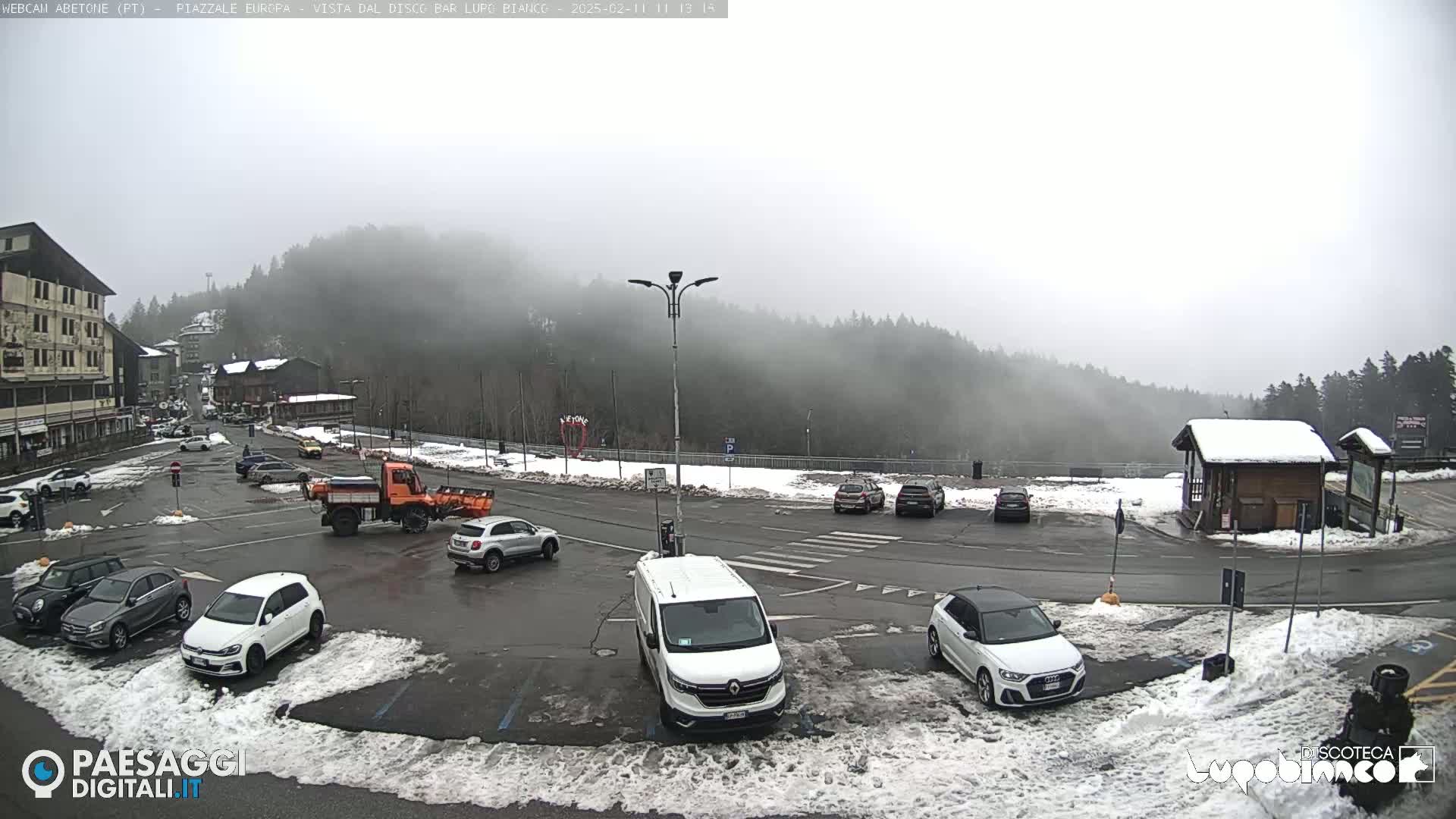 A foggy parking lot with several cars and a snow plow, surrounded by snow-covered ground and a wooded hillside in the background.