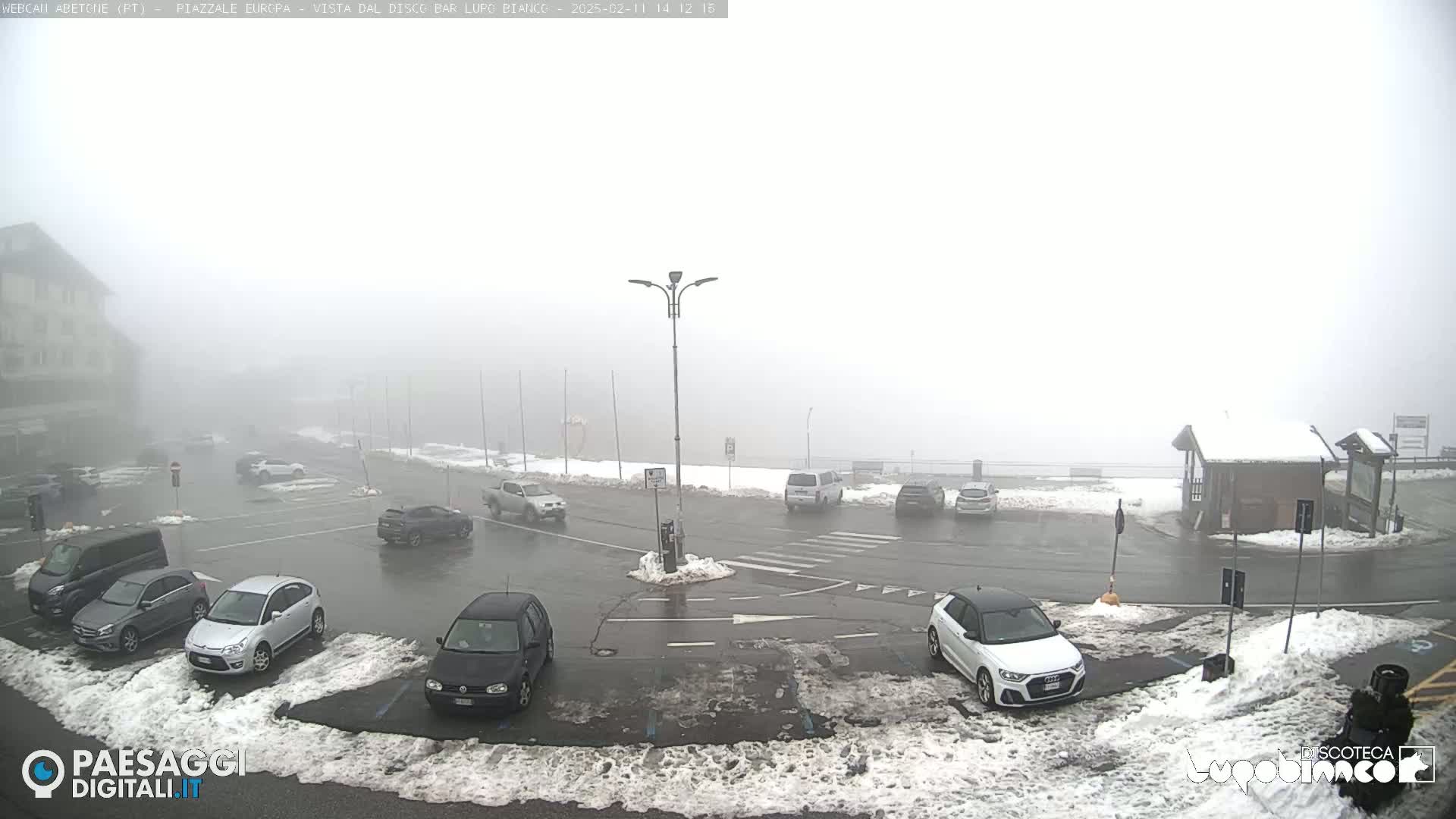 A foggy parking lot with several cars, partially covered in snow, is situated near a building.