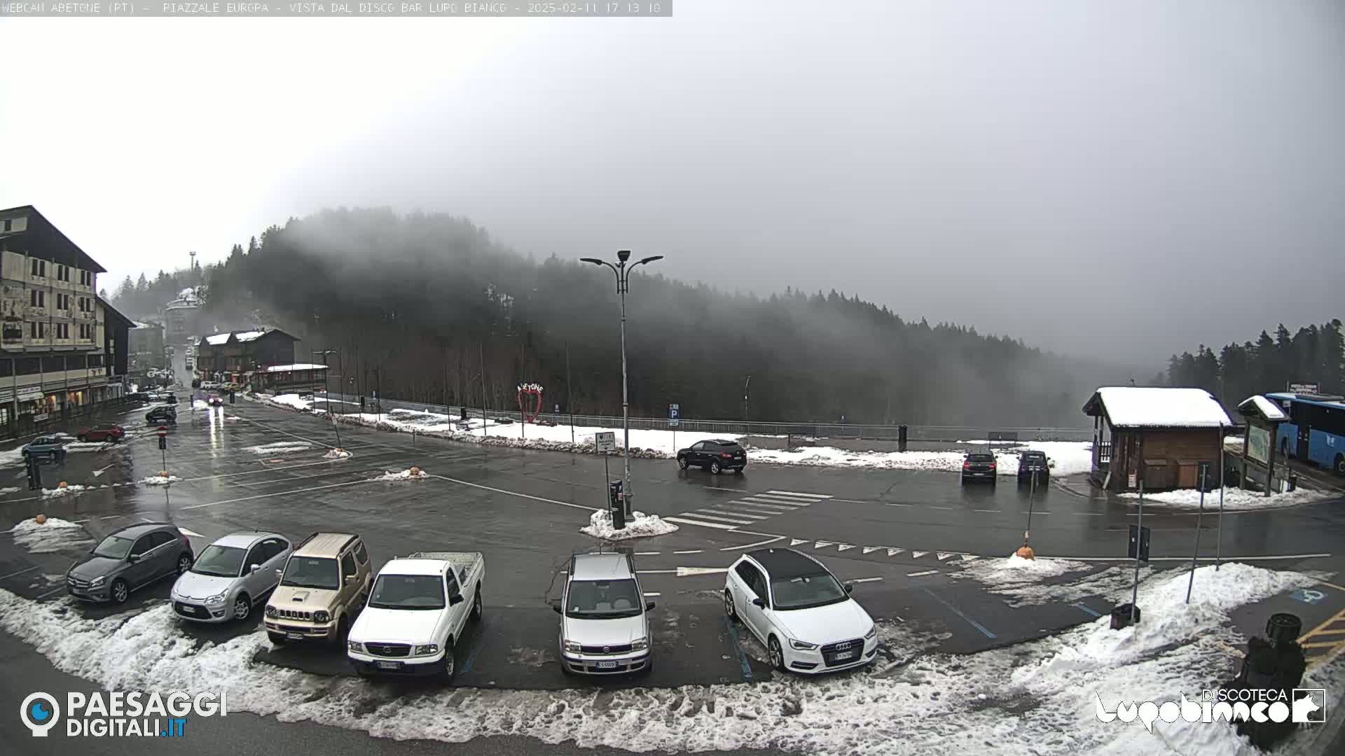 A mostly empty parking lot with several cars is situated in front of a building and forested hills shrouded in fog on a snowy, overcast day.