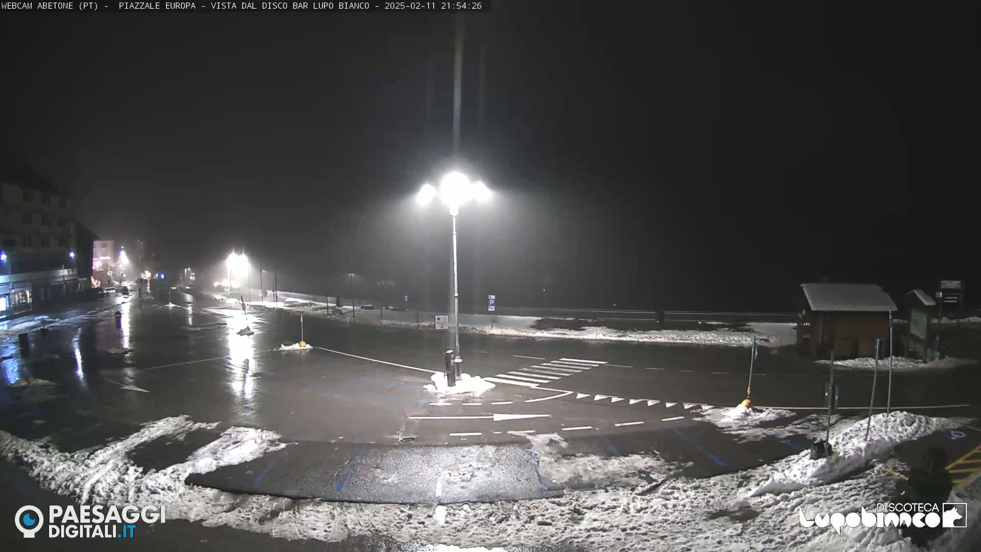 A nighttime scene of a mostly empty paved area with some snow on the ground and wet pavement, illuminated by streetlights under a light rain or snow.