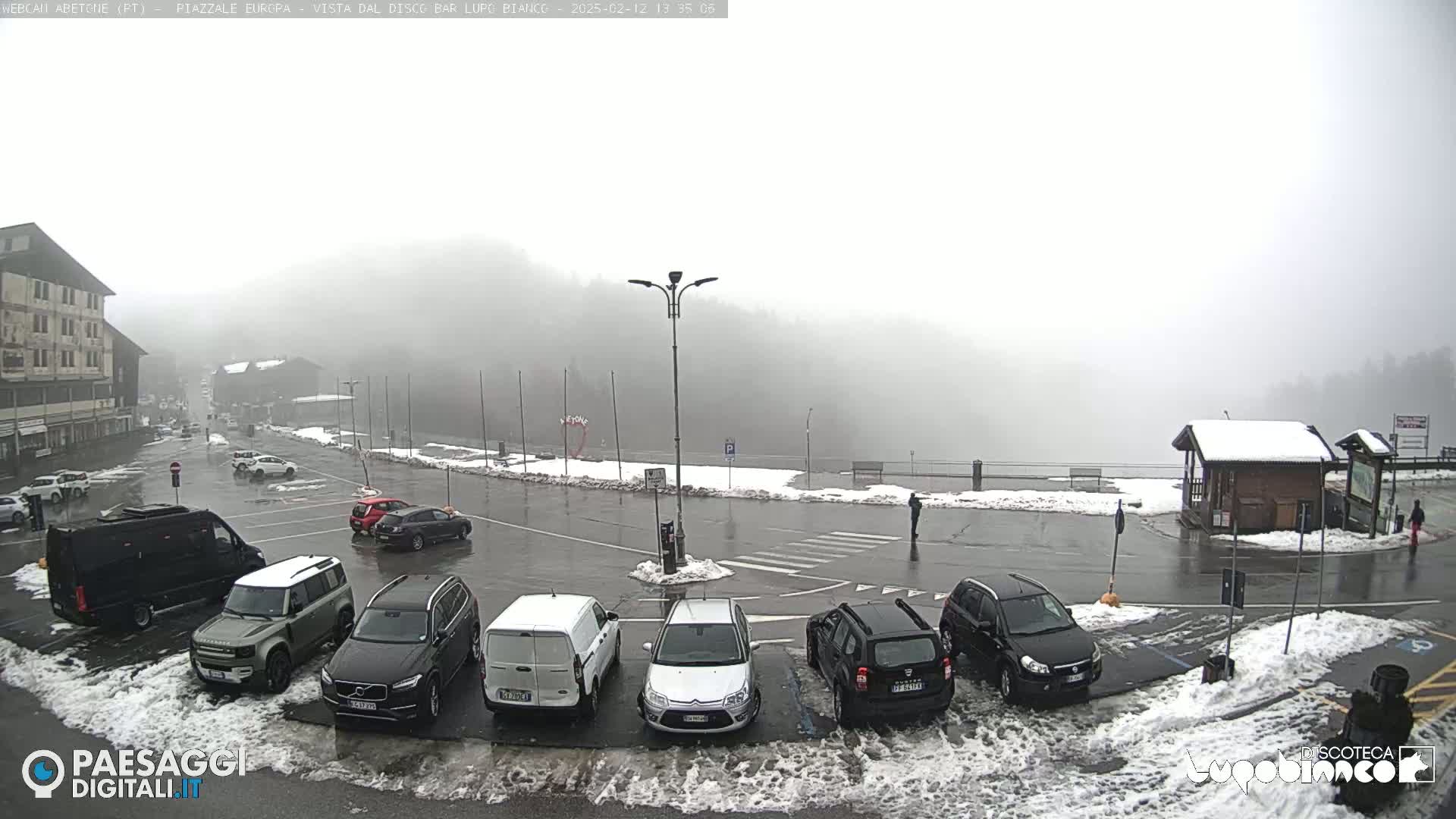A foggy parking lot with several cars and a building in the background, some patches of snow are visible on the ground.