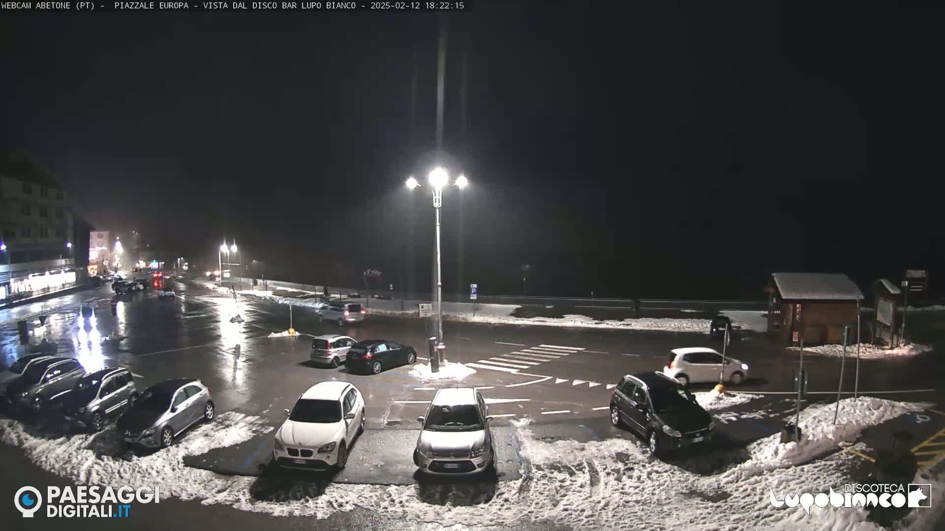 A nighttime parking lot with several cars parked in it, some patches of snow on the ground, and wet pavement under streetlights.