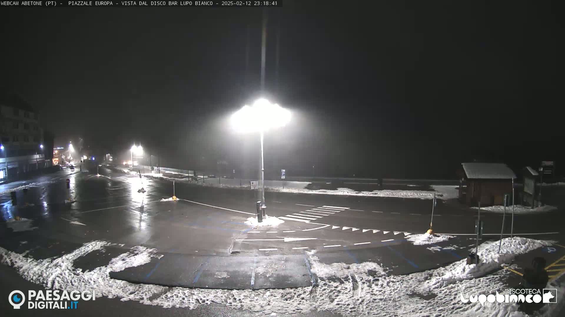 A nighttime, snow-dusted parking lot and roadway are illuminated by bright streetlights under foggy conditions.