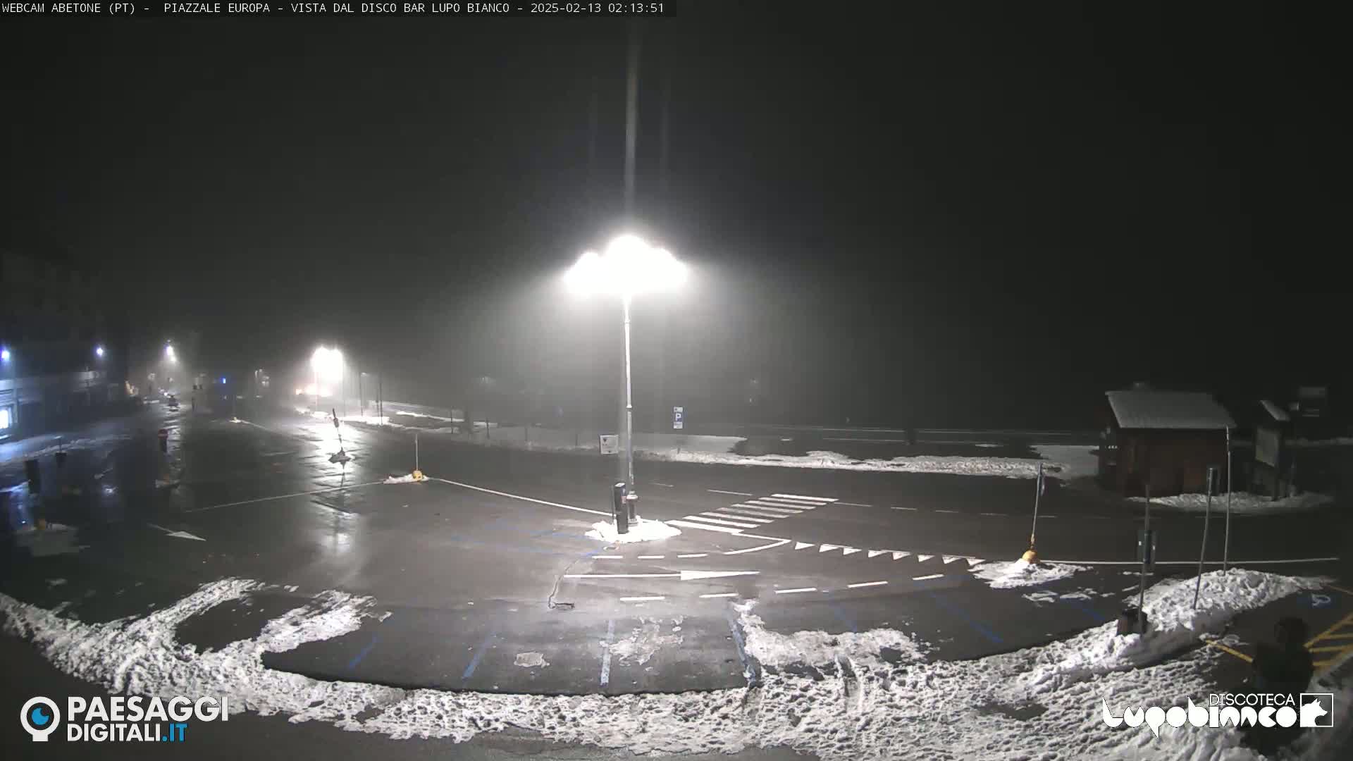 A nighttime, foggy view of a mostly empty parking lot with some snow on the ground and a bright streetlight illuminating the scene.