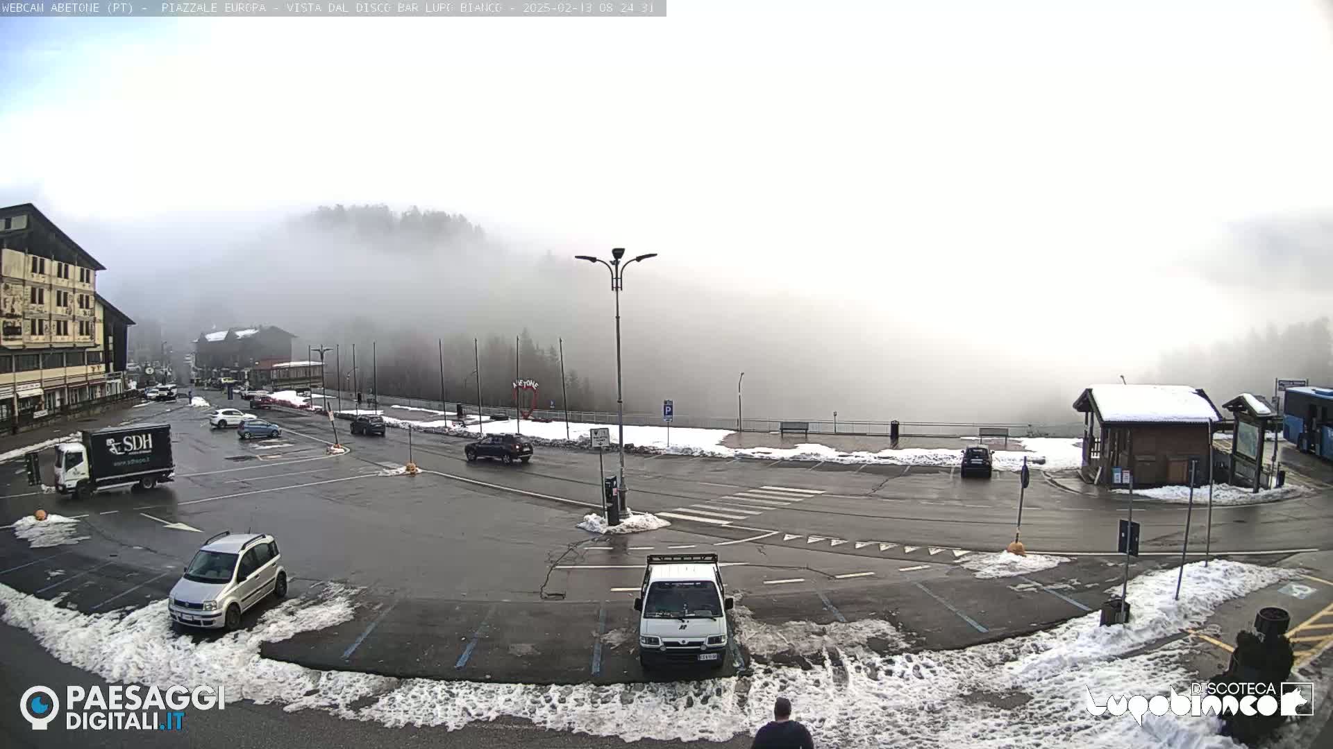 A foggy parking lot with several cars and trucks, surrounded by snow-covered areas and buildings, is shown with a building in the background partially obscured by fog.