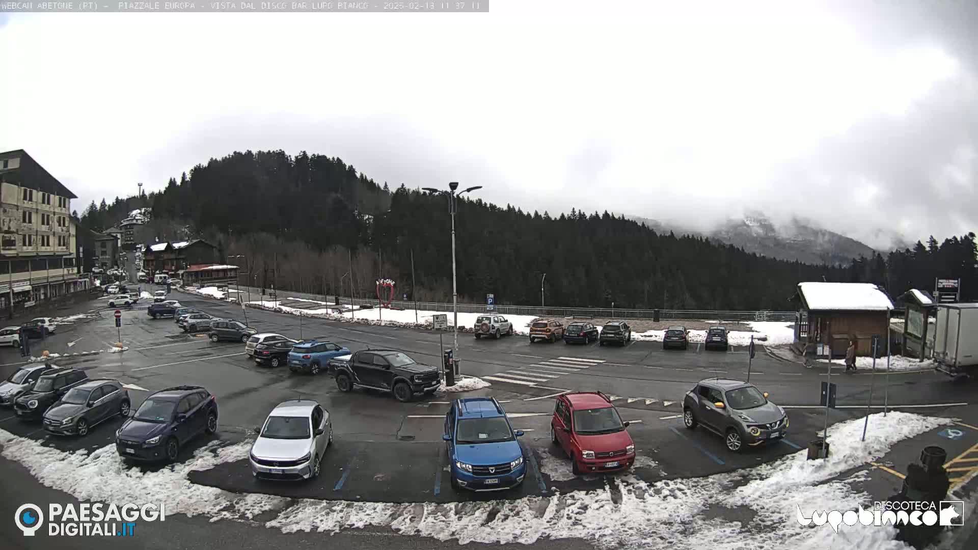 A parking lot with numerous cars is situated in front of a building, with a snow-covered landscape and overcast skies in the background.
