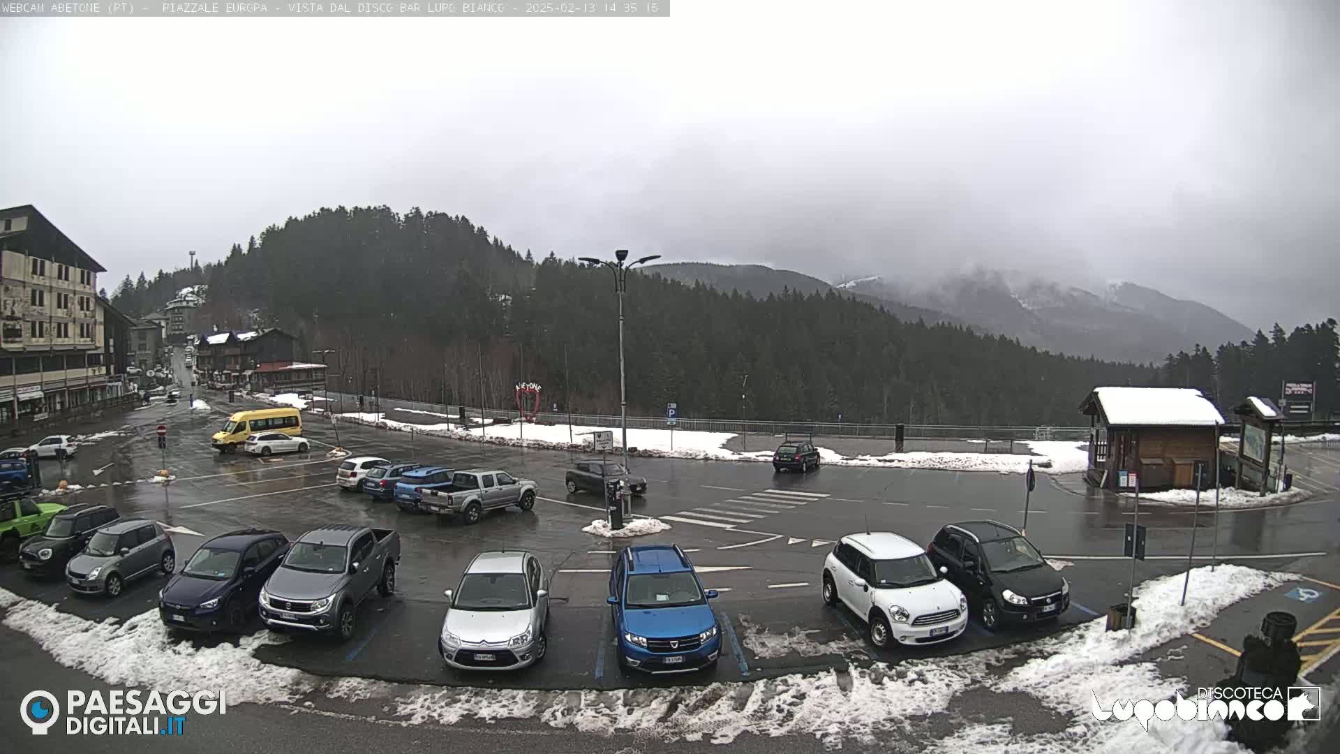 A wet parking lot with several cars, a van, and a small building, surrounded by snow and overlooking a mountain range under an overcast sky.