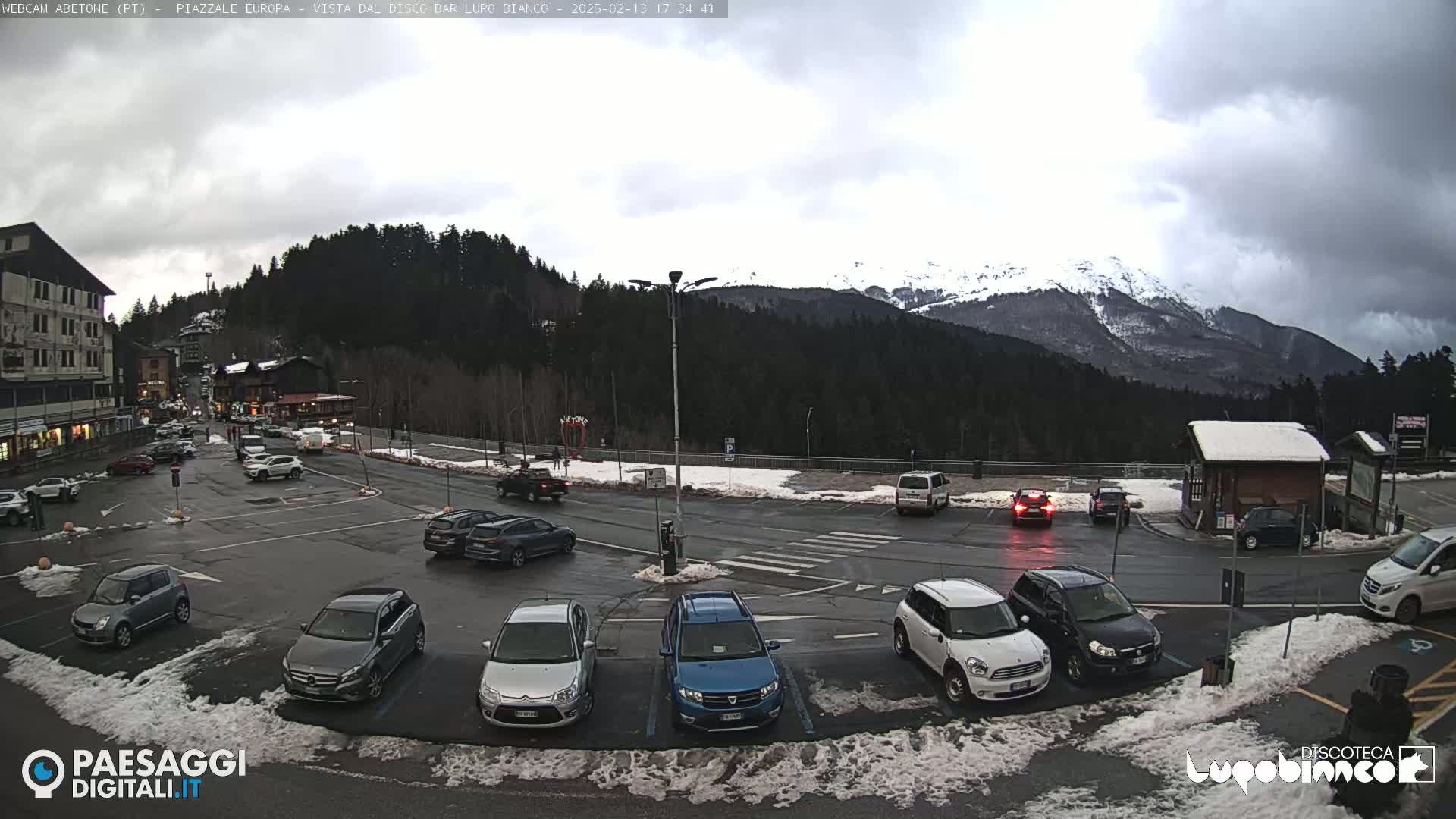 A parking lot with several cars is situated in front of a building and a mountain range under an overcast sky.