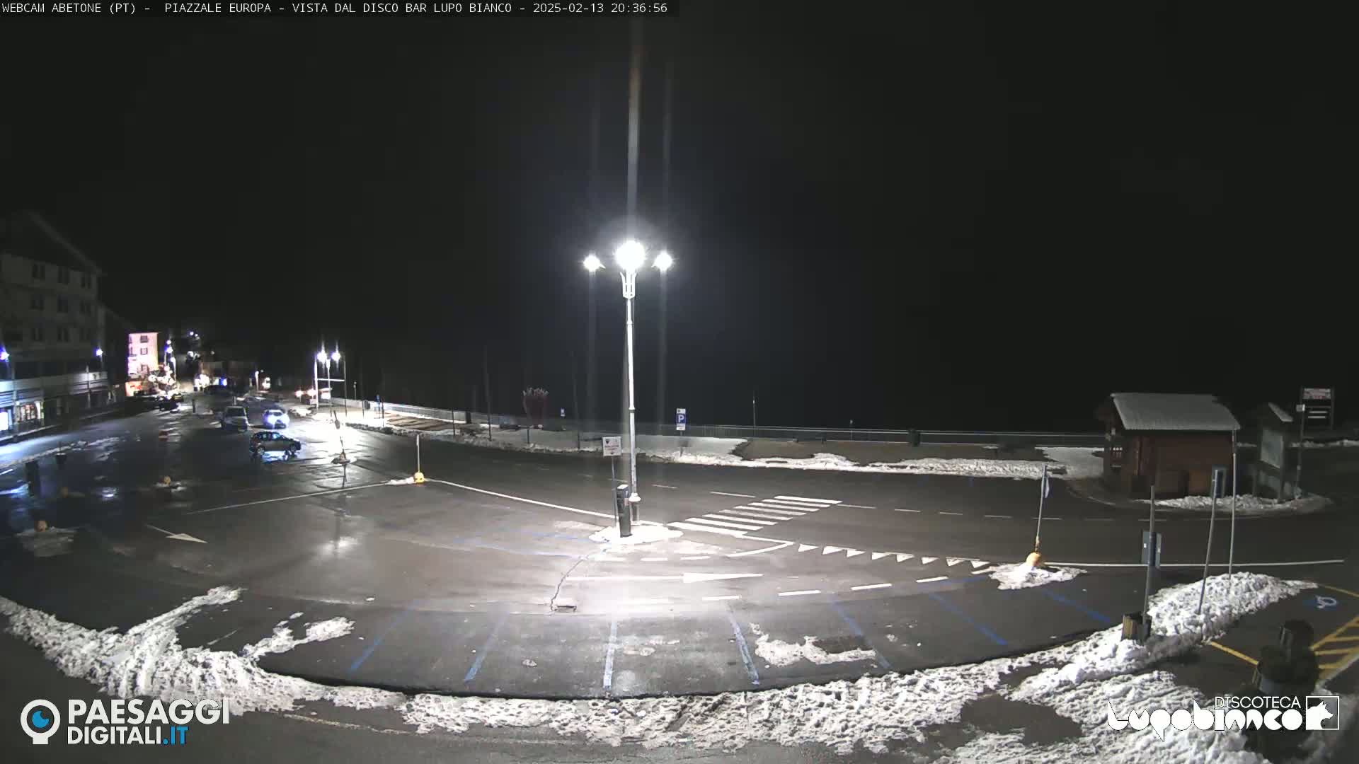 A mostly empty, snow-dusted parking lot and roadway are illuminated at night by streetlights, with a few cars visible and a building in the background.
