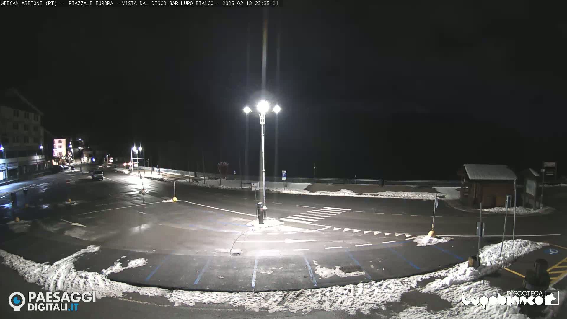 An empty, snow-dusted parking lot at night, illuminated by streetlights, is bordered by buildings and a road.