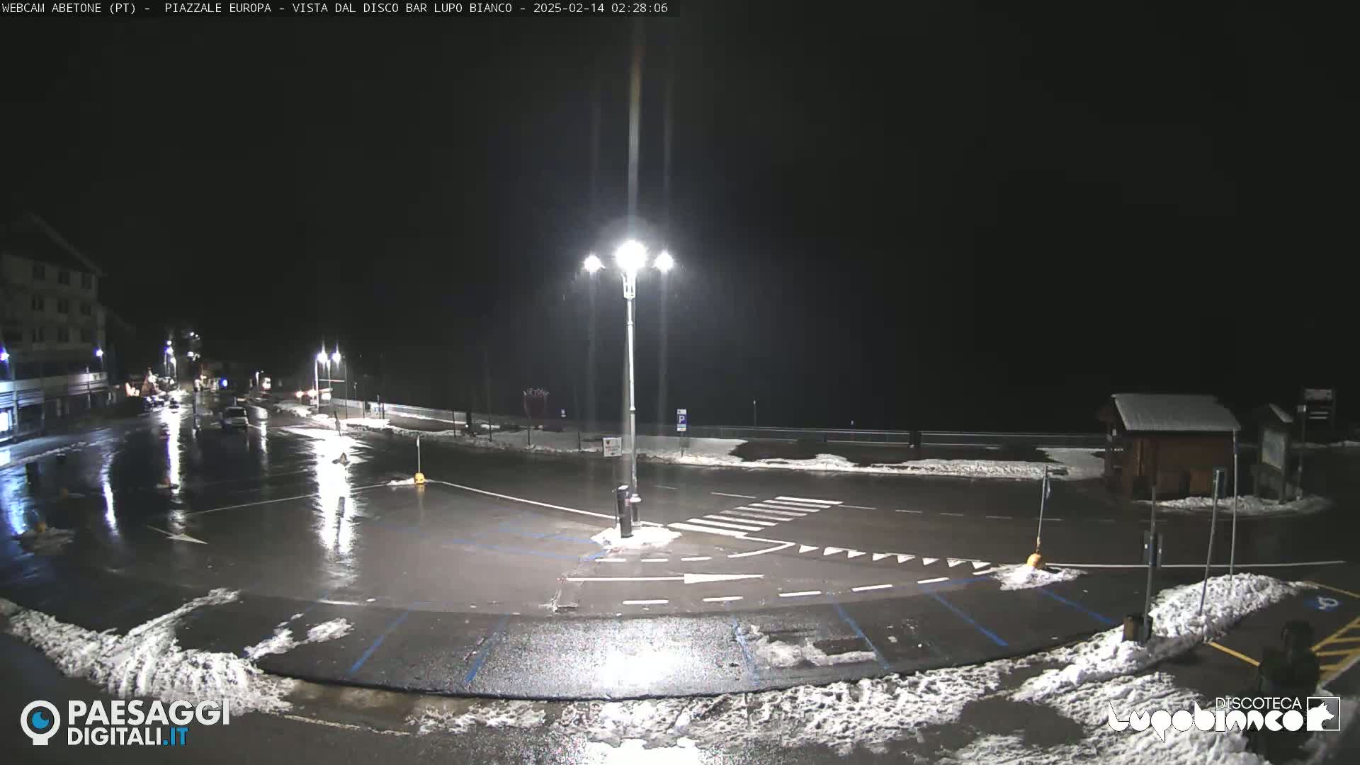 A nighttime, wet parking lot with some snow on the ground and a few cars is illuminated by streetlights.